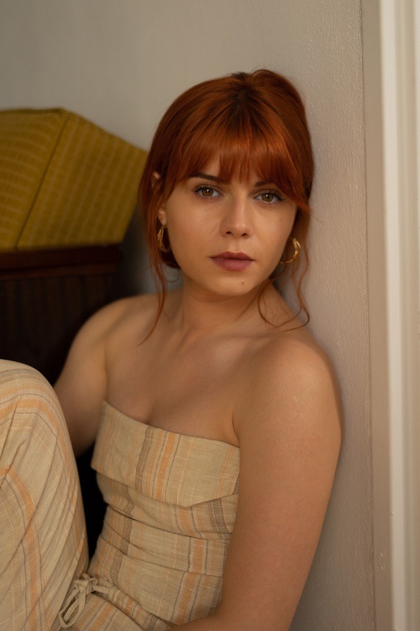 Portret kobiety o rudych włosach z grzywką, w beżowym kombinezonie w paski, opierającej się o ścianę, z kolczykami kołami.