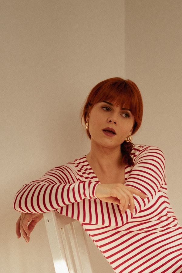 Portret kobiety z rudymi włosami w bluzce w czerwono-białe paski, opierającej się o białe krzesło, z widocznymi złotymi kolczykami kołami.