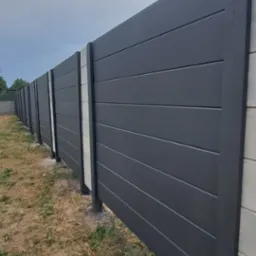 Nowoczesne ogrodzenie z paneli w kolorze antracytowym, z betonowymi słupkami, widoczne z boku, z suchą trawą u podstawy.