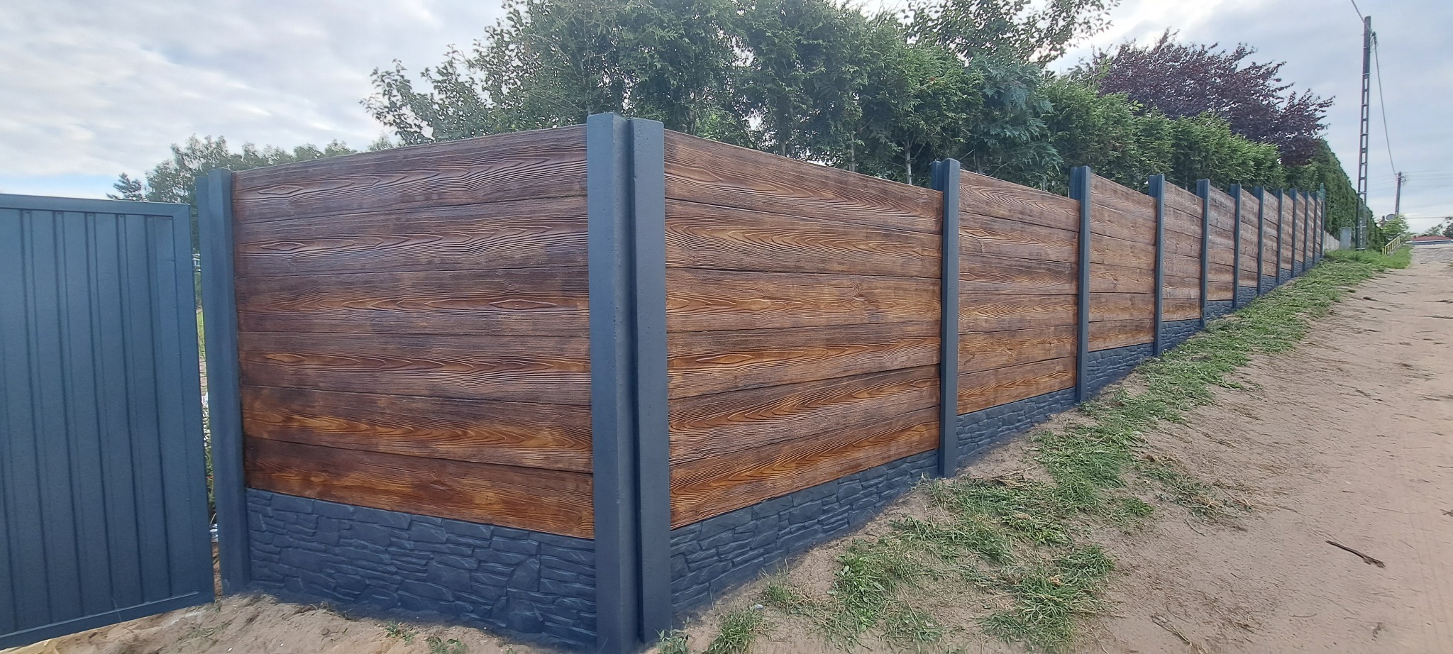 Długi, brązowy płot z imitacją drewna i szarymi słupkami oraz podmurówką, biegnący wzdłuż piaszczystej drogi, z zielenią w tle.