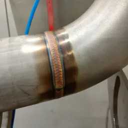 Spawany element rury ze stali nierdzewnej z widocznymi kolorami spawu, niebieski przewód w tle.