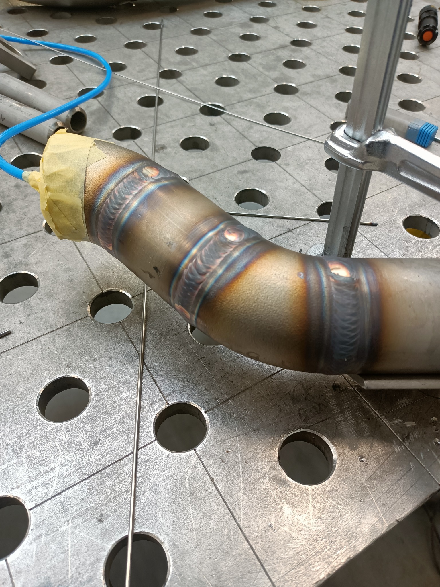 Spoina na zagiętej rurze metalowej, widoczne ślady ciepła, zabezpieczona taśmą, leżąca na perforowanym stole spawalniczym.