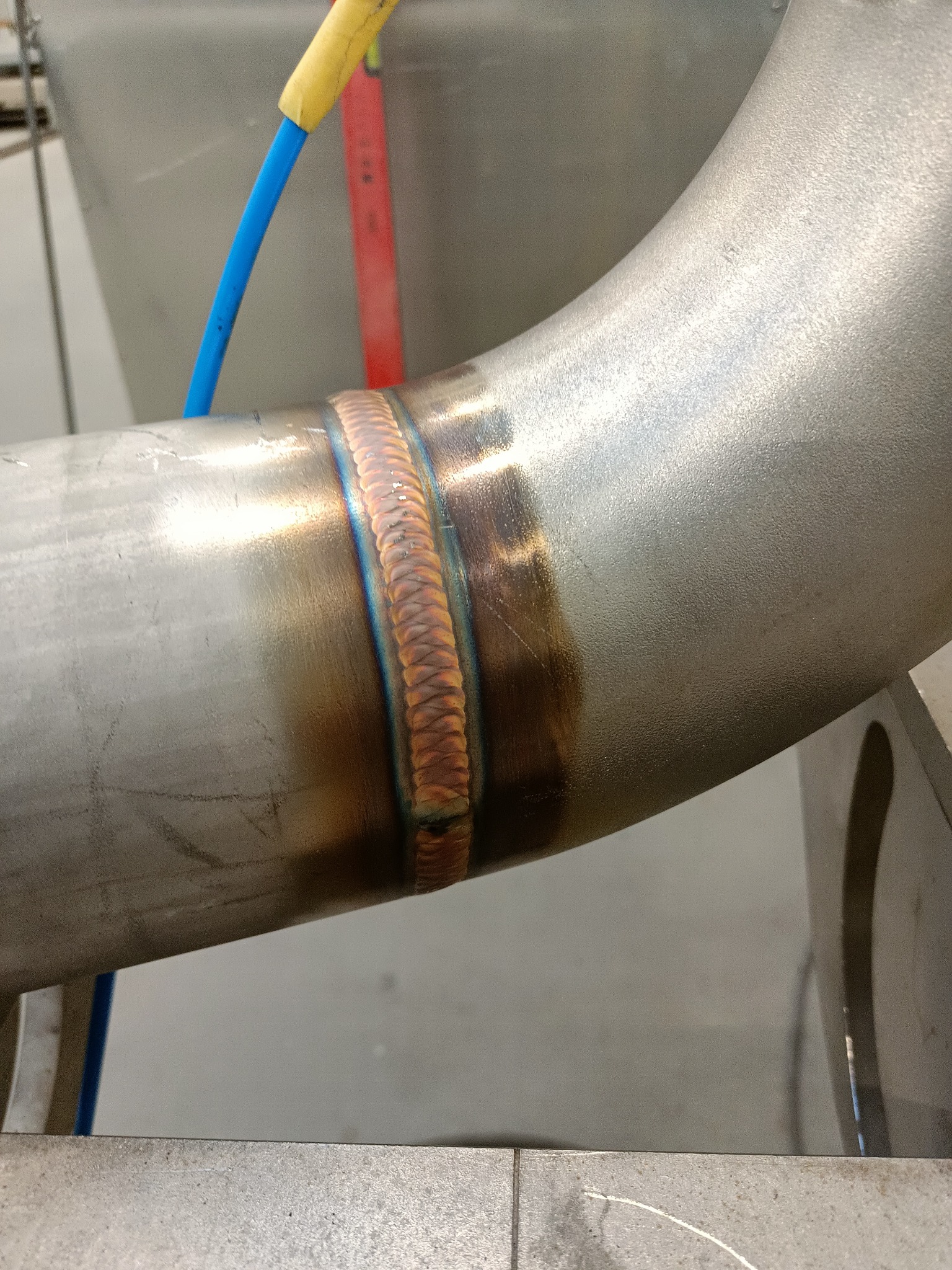 Spawany element rury ze stali nierdzewnej z widocznymi kolorami spawu, niebieski przewód w tle.