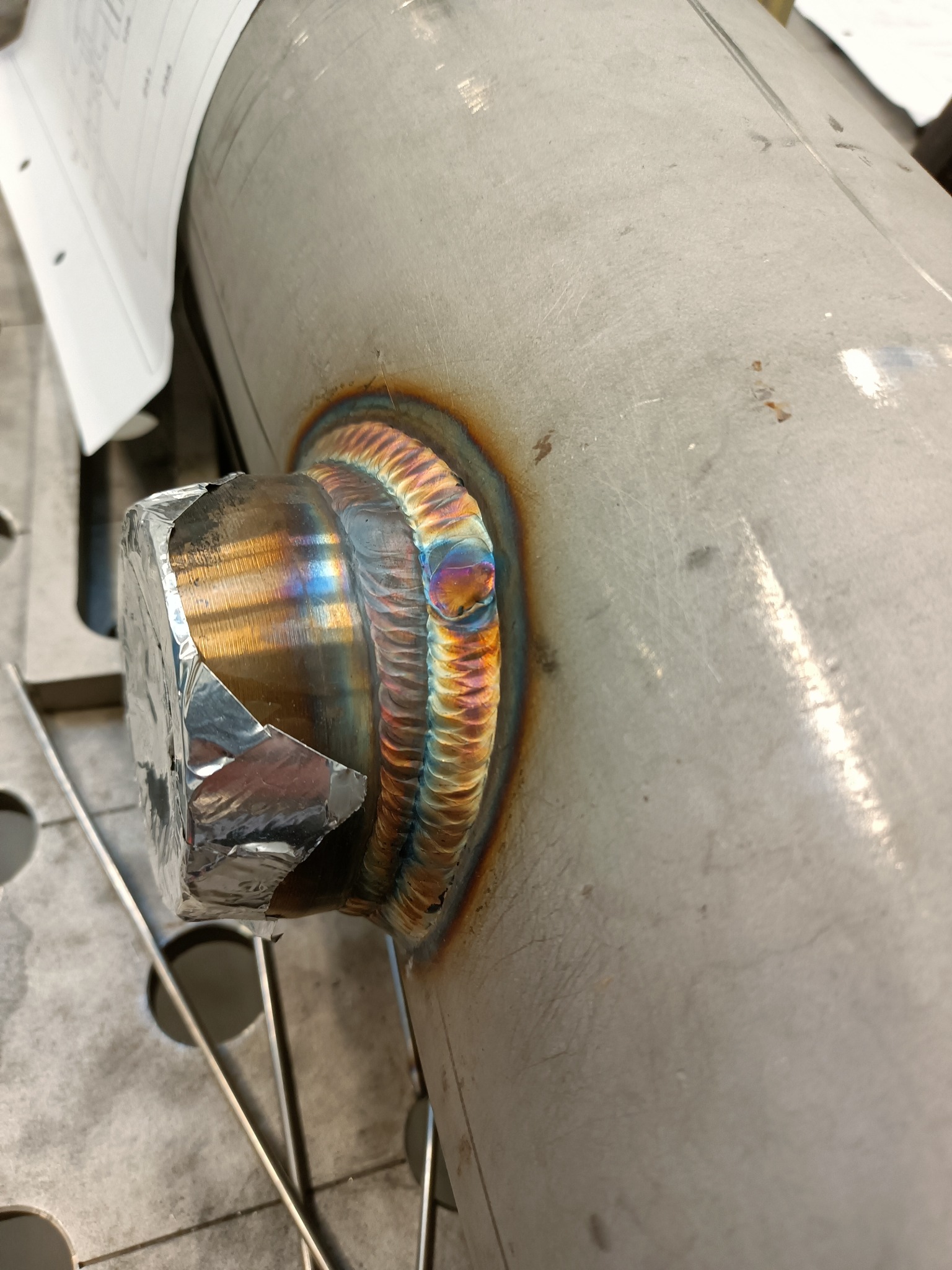 Złącze spawane rury ze stali nierdzewnej, widoczne tęczowe przebarwienia spoiny, zabezpieczone folią aluminiową, na tle metalowego stołu spawalniczego.