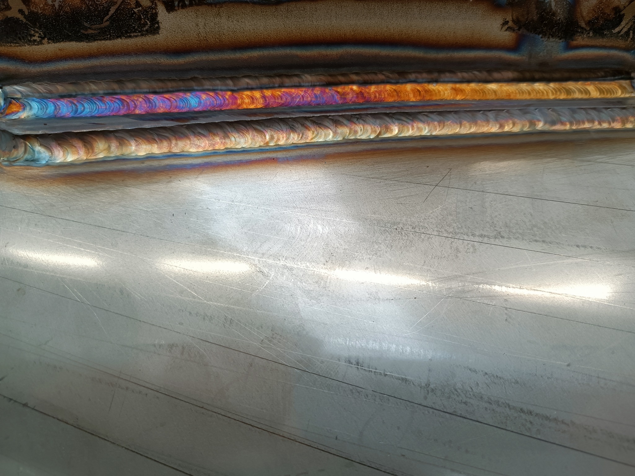 Szczegółowe zbliżenie trzech równoległych spoin na metalowej powierzchni, z wyraźnymi śladami obróbki i tęczowymi przebarwieniami od ciepła.