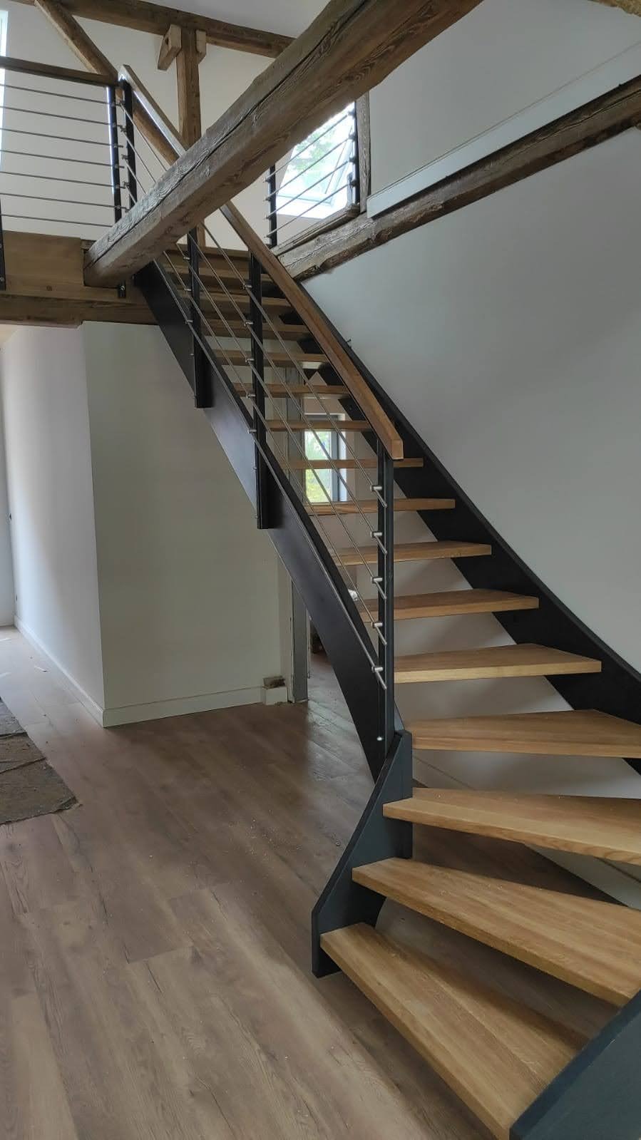 Nowoczesne schody z drewnianymi stopniami, metalową konstrukcją i balustradą z cienkich prętów, w jasnym wnętrzu z drewnianymi belkami stropowymi.