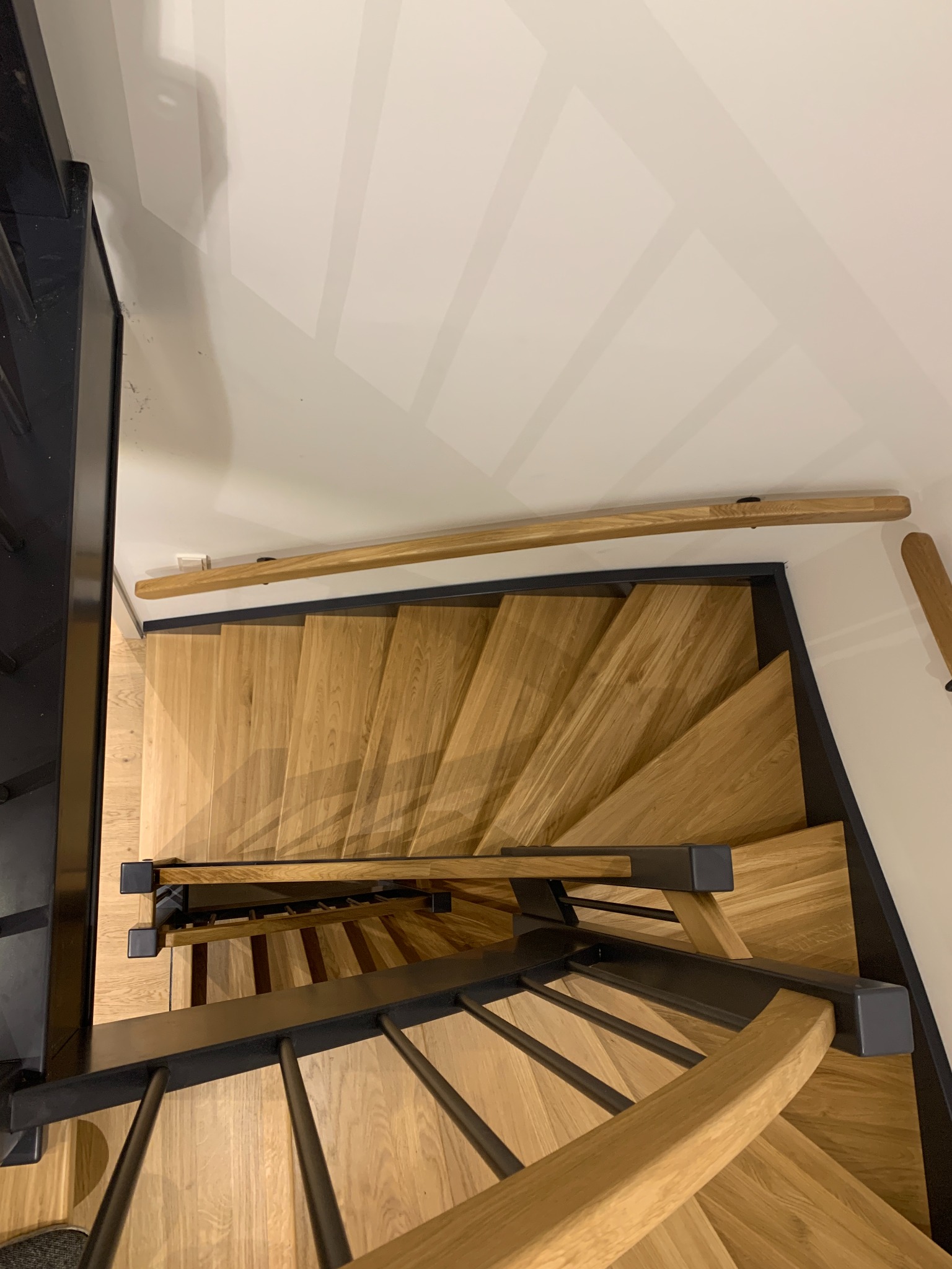 Widok z góry na kręte schody z jasnego drewna z czarnymi metalowymi balustradami, zamontowane przy białej ścianie z szarymi paskami dekoracyjnymi.