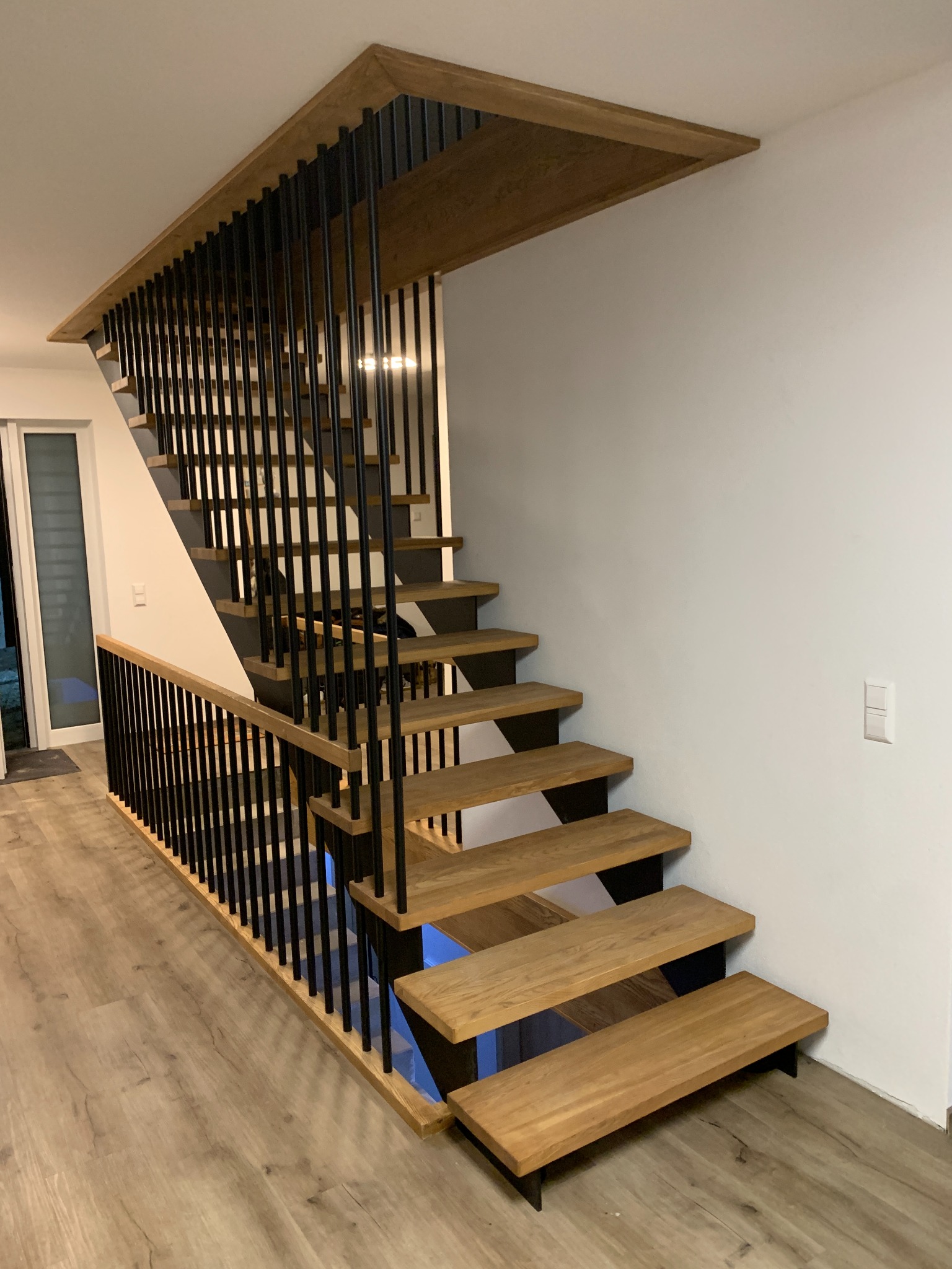 Nowoczesne schody drewniane z czarnymi metalowymi balustradami pionowymi, dębowe stopnie i podświetlenie LED pod stopniami, widok z dołu na jasnym parkiecie.