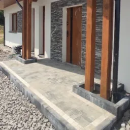 Nowoczesny podjazd z kostki brukowej w odcieniach szarości, obramowany jasnymi kostkami, prowadzący do wejścia domu z drewnianymi słupami i elewacją z kamienia dekoracyjnego.