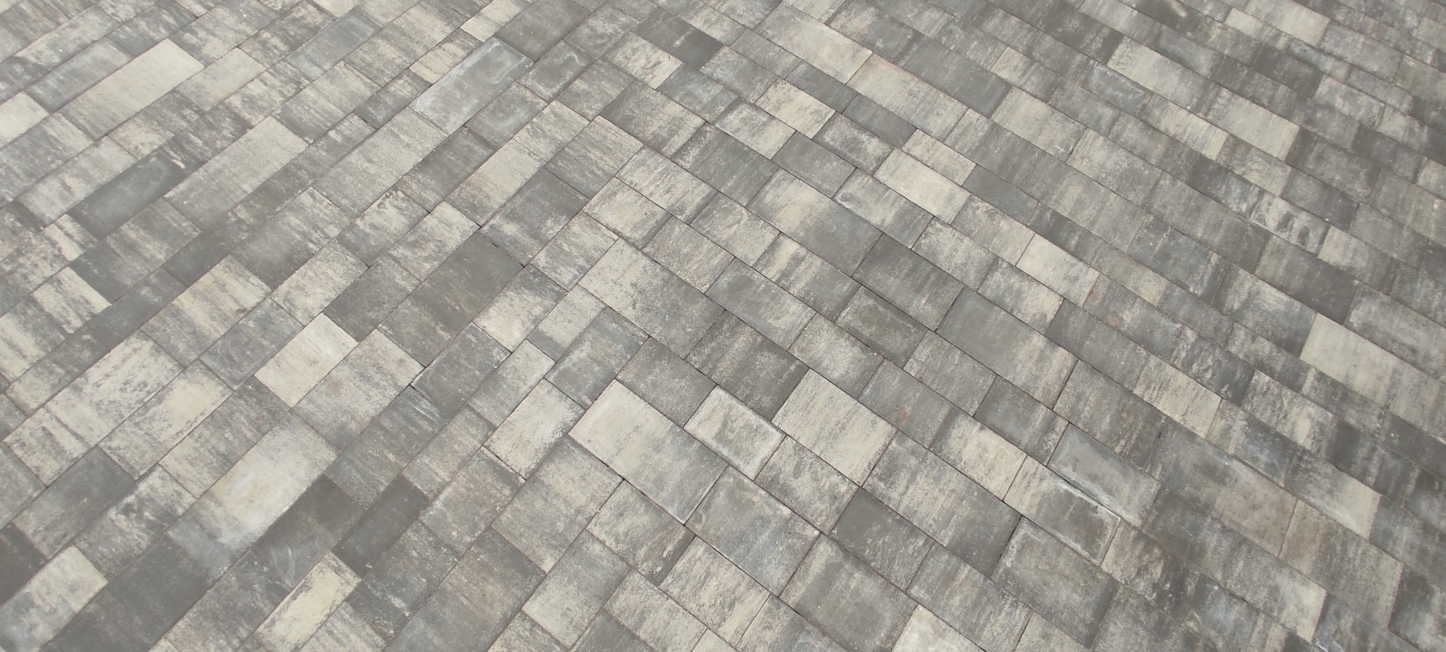 Ułożona szara kostka brukowa w jodełkę, widoczna z góry, z delikatną teksturą i subtelnymi różnicami w odcieniach szarości na poszczególnych elementach.