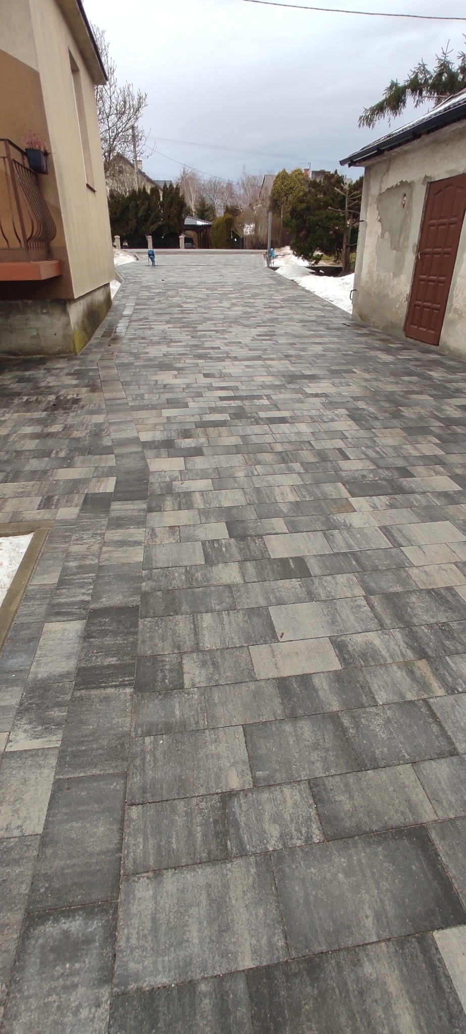 Szeroki podjazd z szarej kostki brukowej ułożonej w prostokątny wzór, z budynkami po bokach i resztkami śniegu na obrzeżach.