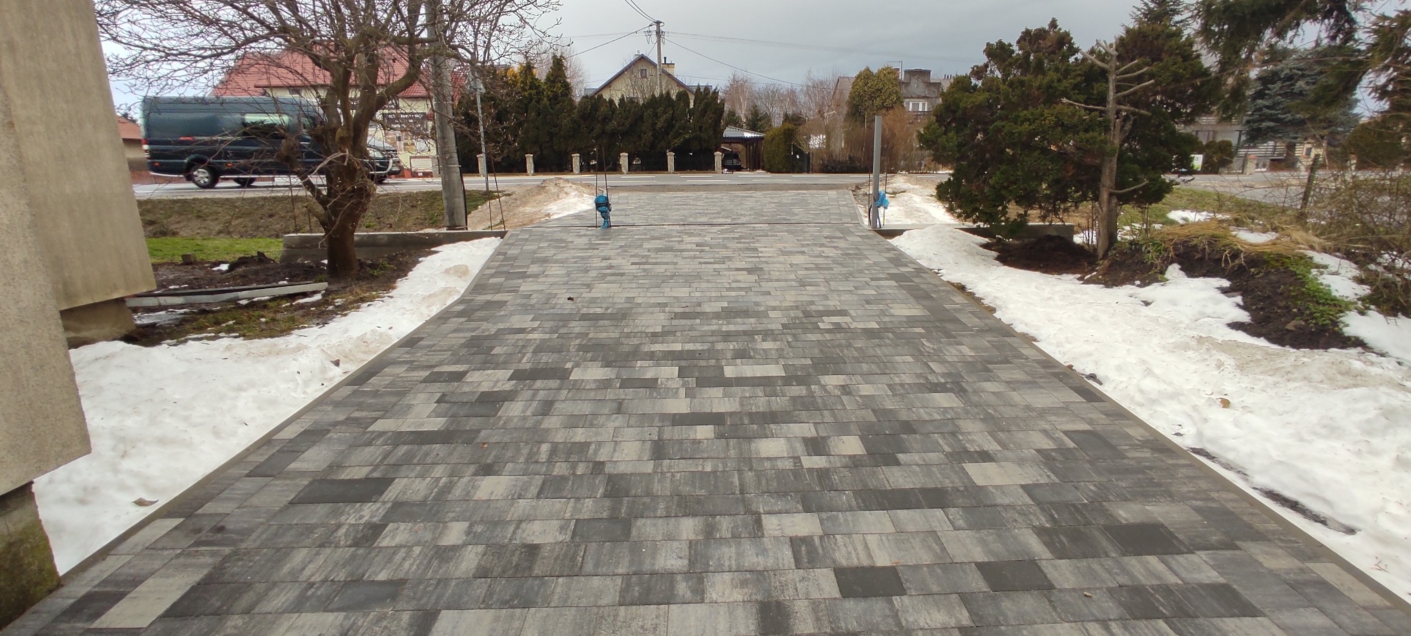 Nowa, szara nawierzchnia z kostki brukowej prowadząca do posesji, z resztkami topniejącego śniegu po bokach i widocznym budynkiem w tle.