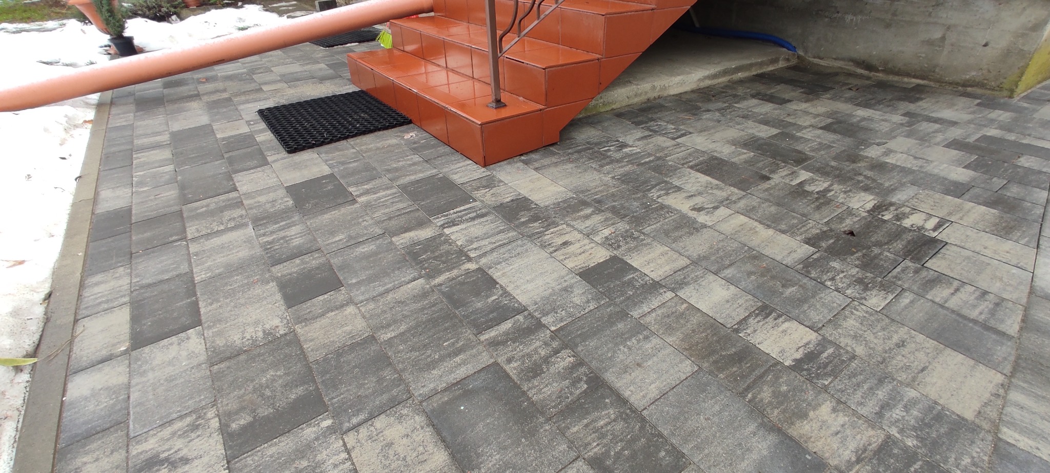 Ułożona szara kostka brukowa prowadząca do pomarańczowych schodów wejściowych z metalową barierką, widoczna wycieraczka i rura spustowa.