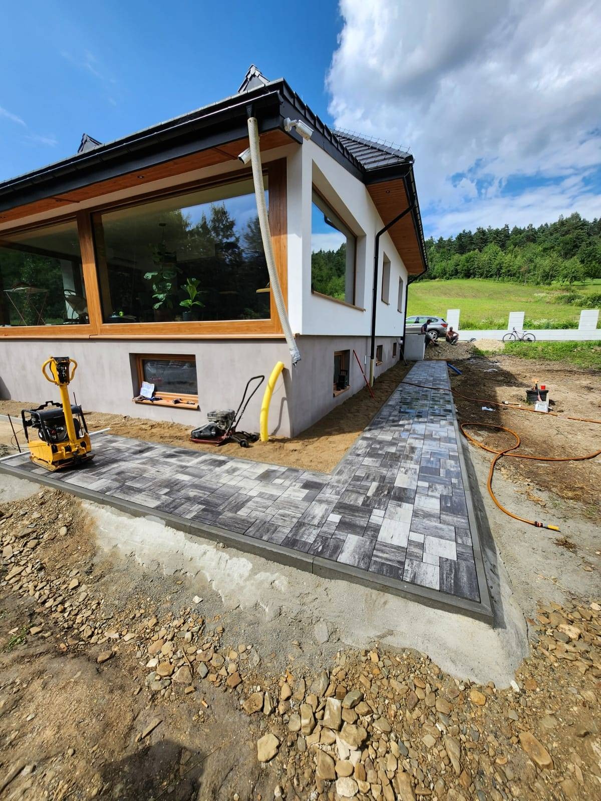 Układanie szarej kostki brukowej wzór melanż na ścieżce wokół domu z widocznymi narzędziami: zagęszczarka, kosiarka, węże.