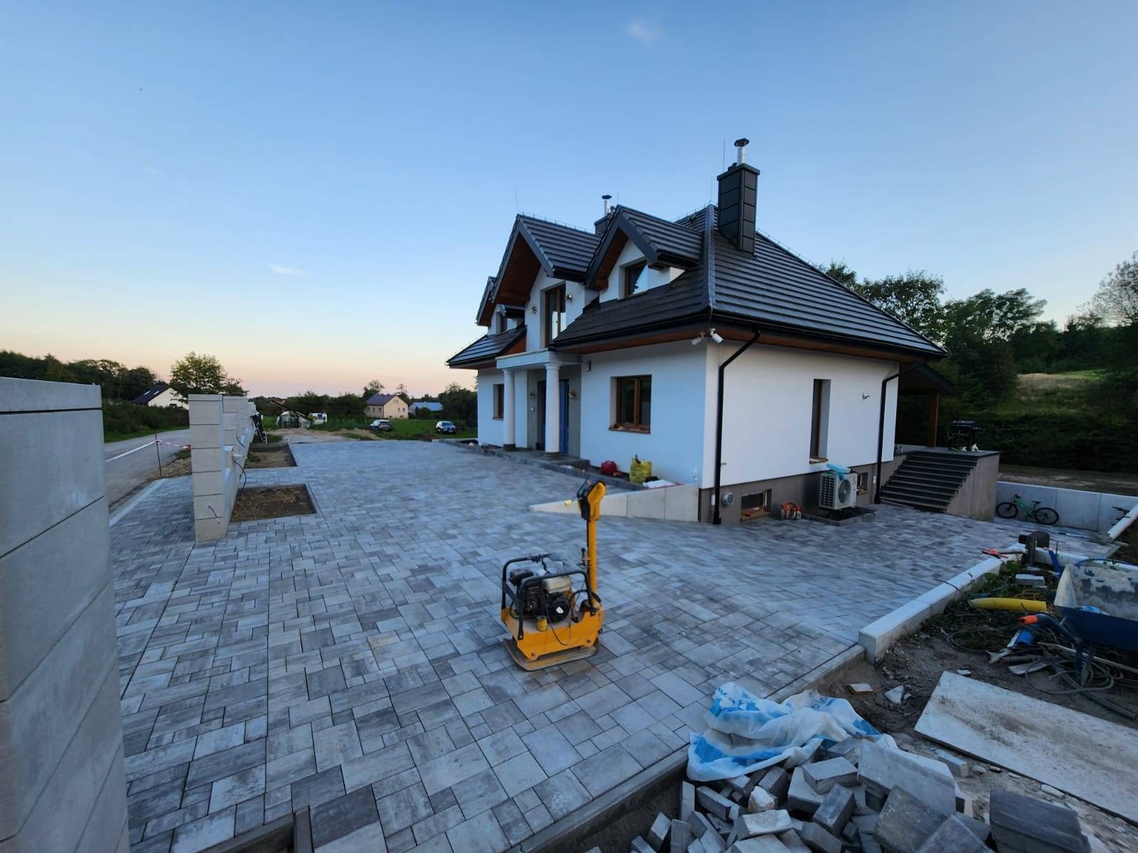 Ułożona szara kostka brukowa na podjeździe przed nowoczesnym domem jednorodzinnym, widoczna zagęszczarka wibracyjna i stos kostki do wykończenia