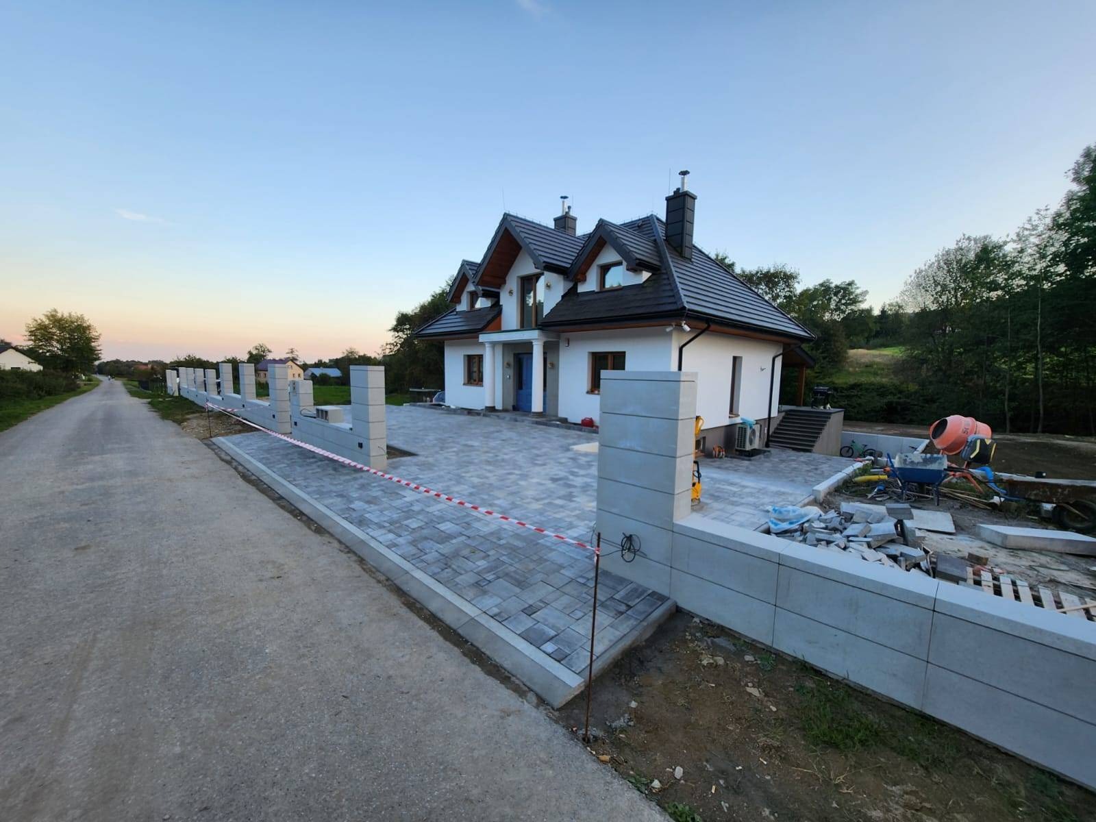 Nowoczesny dom jednorodzinny z szarą kostką brukową na podjeździe i chodniku, ogrodzenie z betonowych elementów, widoczna betoniarka i narzędzia budowlane w tle.