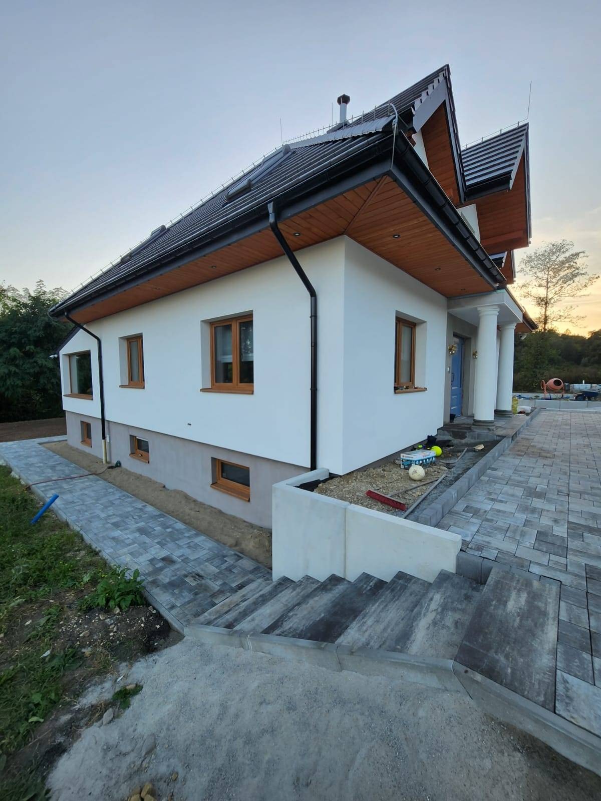 Nowoczesny dom jednorodzinny z wykończoną elewacją, brukowanym chodnikiem i schodami z kostki w odcieniach szarości, widoczne narzędzia i materiały budowlane.