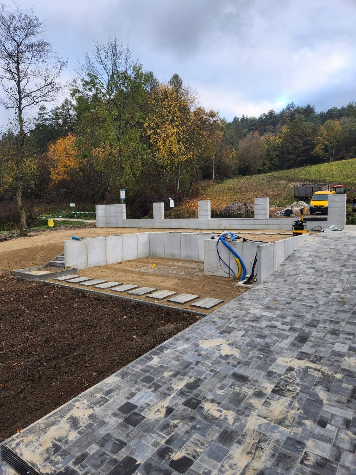 Fundamenty domu z betonu komórkowego, częściowo zasypane piaskiem, z ułożonym fragmentem chodnika z szarej kostki brukowej i przygotowanym terenem pod trawnik, w tle las i żółty samochód dostawczy.