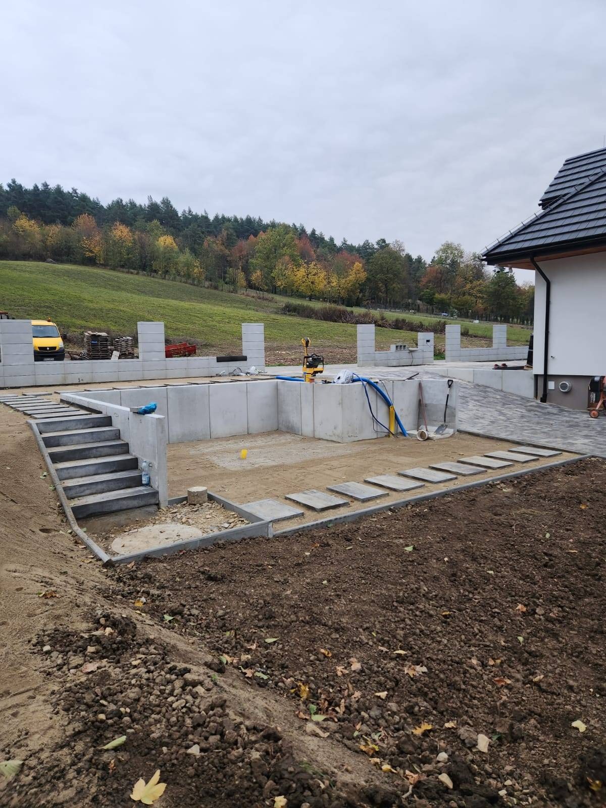 Budowa podjazdu i schodów z kostki betonowej w otoczeniu ziemi, widoczny żółty samochód dostawczy i jesienny krajobraz w tle.