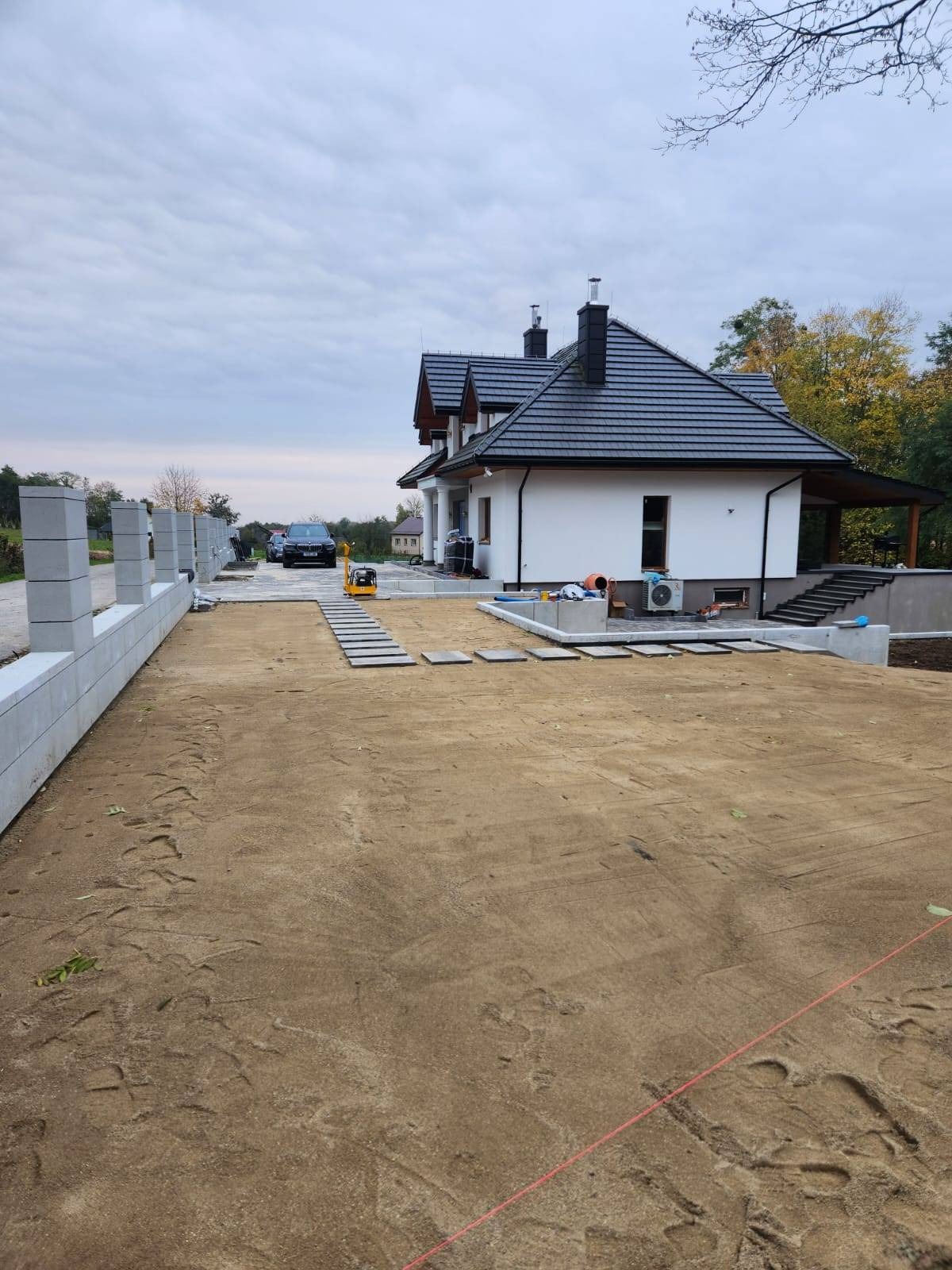 Przygotowany teren pod budowę podjazdu, widoczne ślady po zagęszczarce, częściowo ułożone płyty chodnikowe, w tle nowoczesny dom jednorodzinny z czarną dachówką i szarym ogrodzeniem z betonowych...