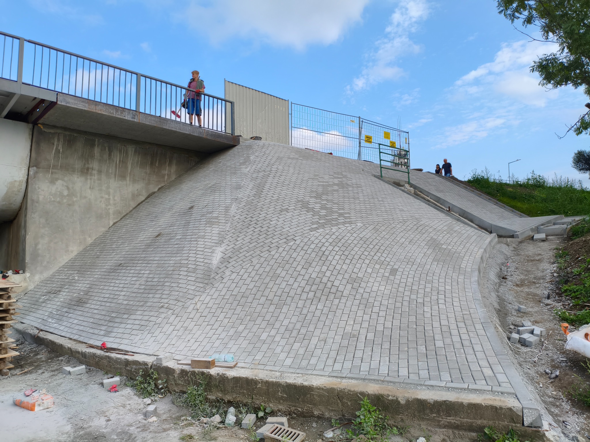 Ukończona nawierzchnia z kostki brukowej na wzniesieniu, widok z dołu. Na górze konstrukcja mostu z osobą. Fragment placu budowy z paletami i materiałami.