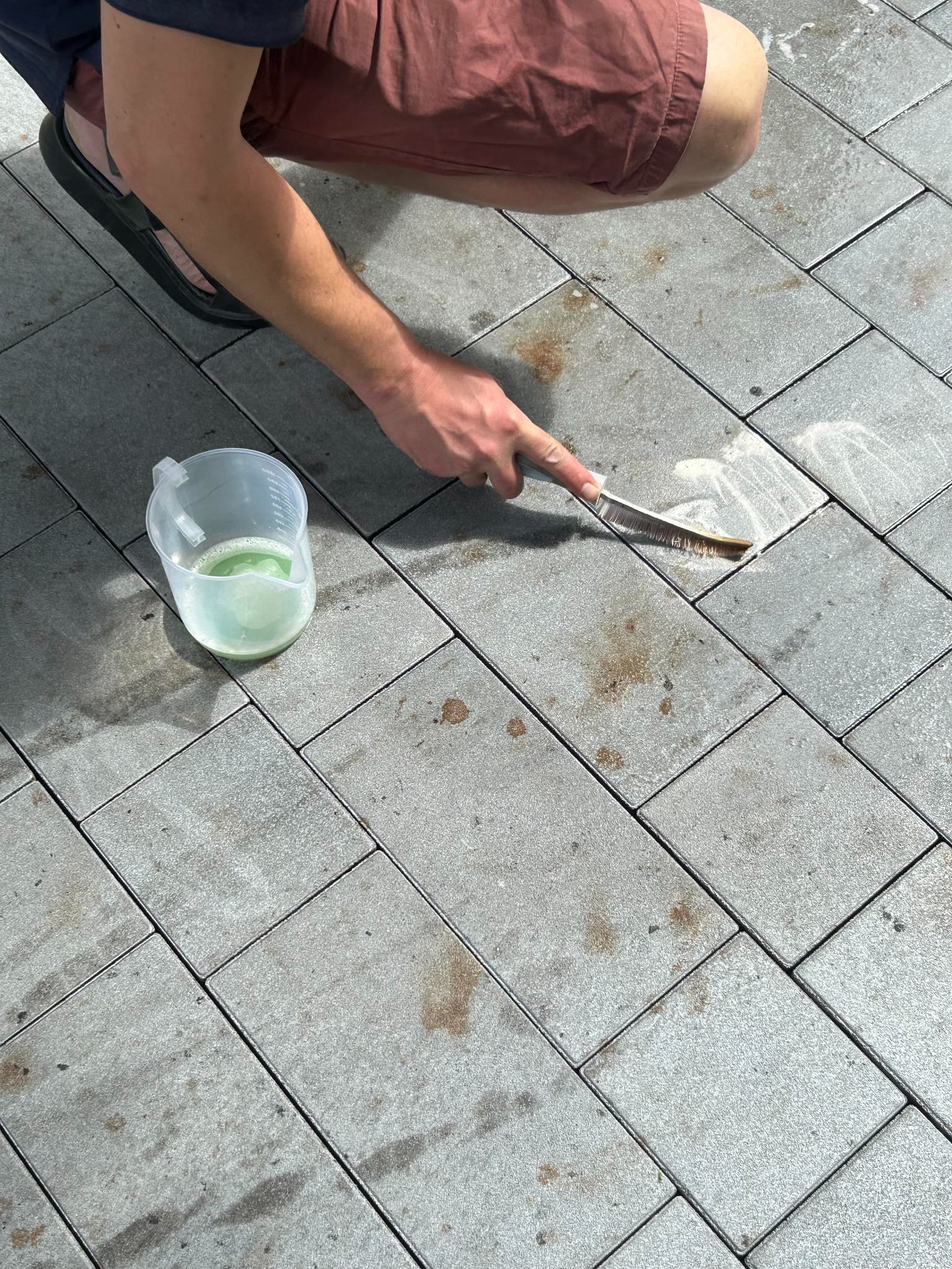 Czyszczenie kostki brukowej szczotką i preparatem z zielonym płynem, widoczne zabrudzenia na kostce przed czyszczeniem.