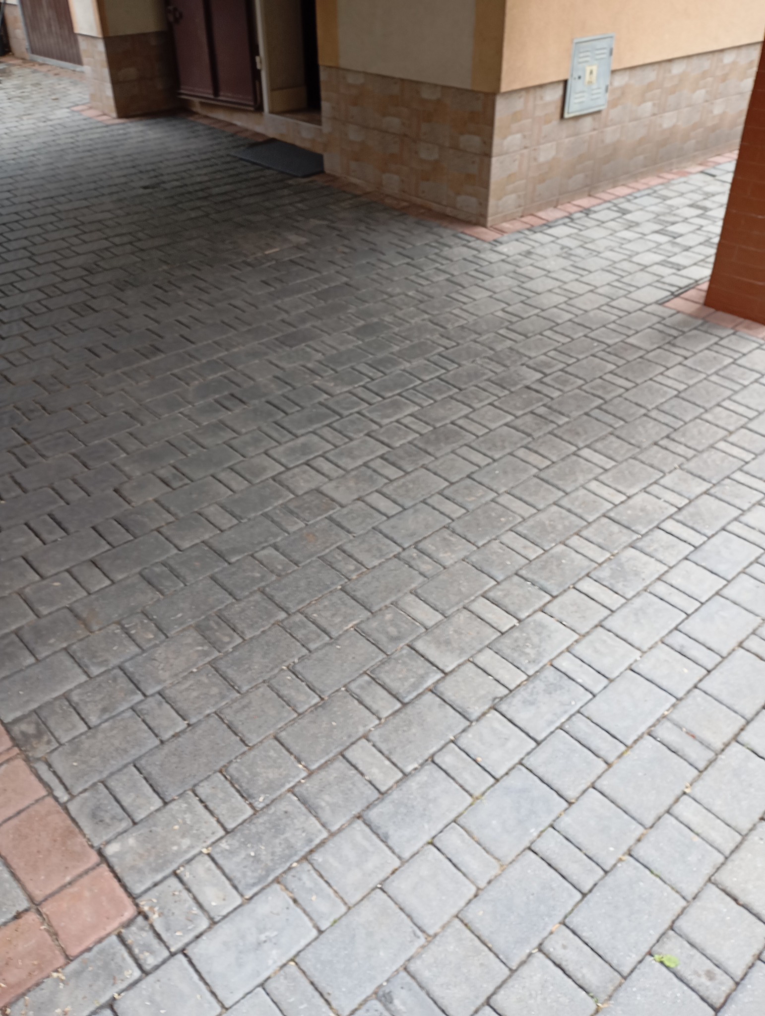 Szara kostka brukowa ułożona pod zadaszeniem budynku, widoczne fragmenty elewacji i drzwi.