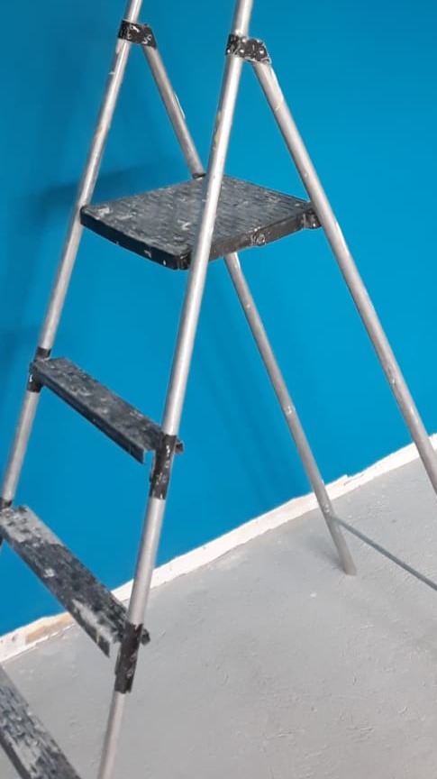 Aluminiowa drabina malarska z zabrudzeniami farby na tle niebieskiej ściany i szarej podłogi.