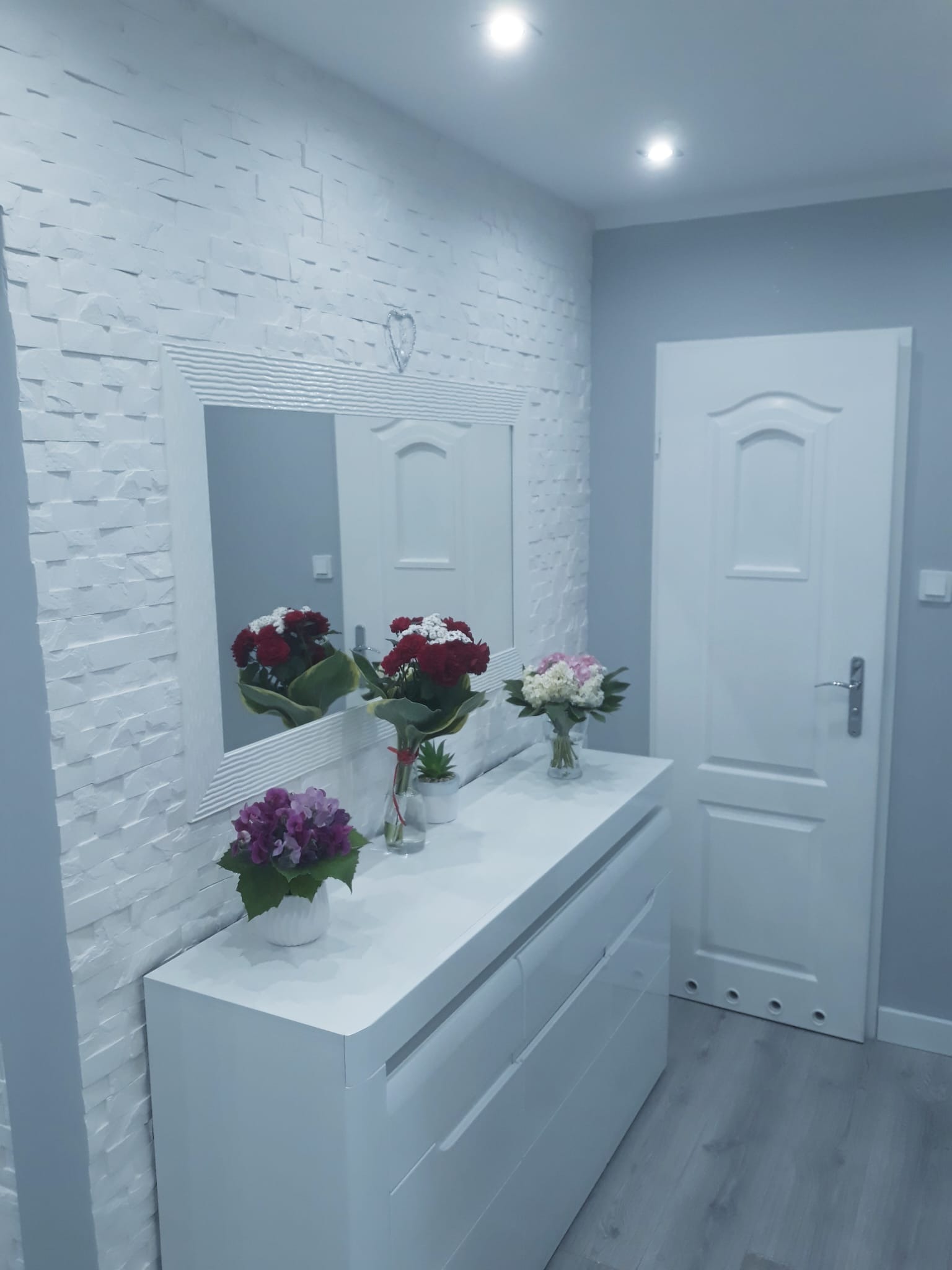 Biała komoda z lustrem w ramie z serduszkiem na ścianie wyłożonej dekoracyjnym białym kamieniem; na komodzie wazony z różnymi kwiatami, białe drzwi w tle.