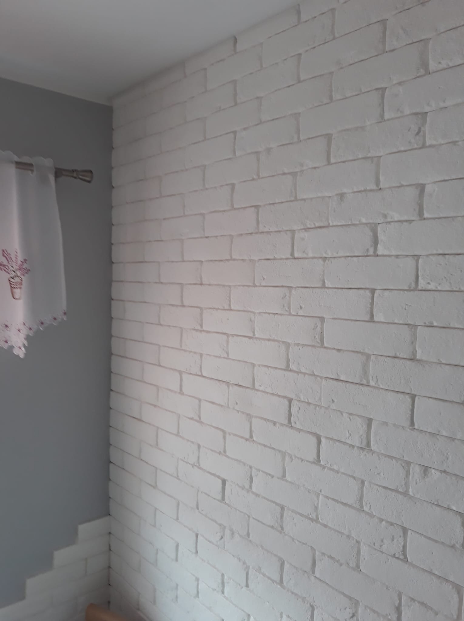 Ściana wykończona białymi, dekoracyjnymi płytkami imitującymi cegłę, fragment zasłony z motywem kwiatowym w rogu.
