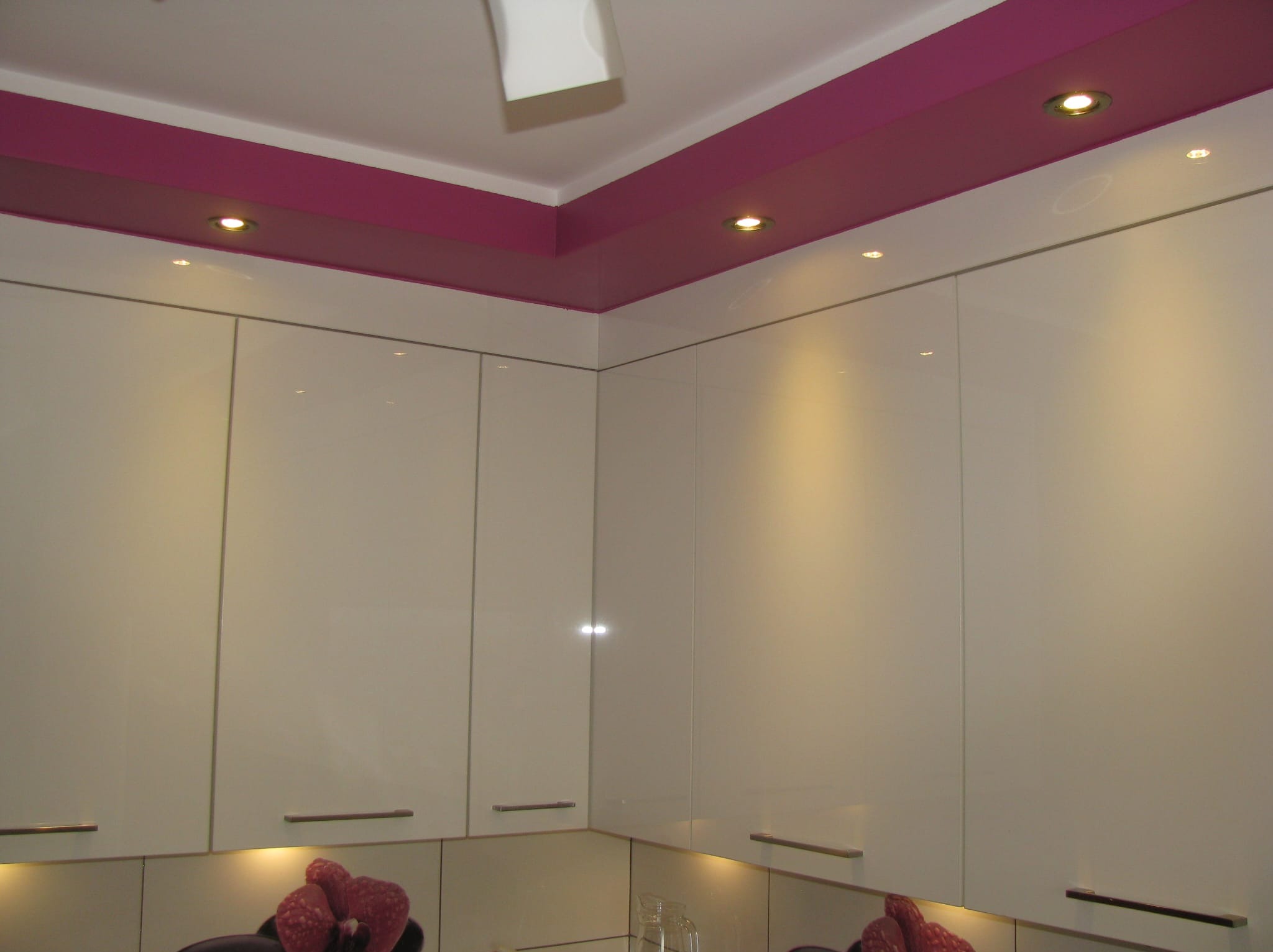 Nowoczesna kuchnia z białymi, błyszczącymi szafkami, podświetlonymi halogenami i purpurowym pasem na suficie.