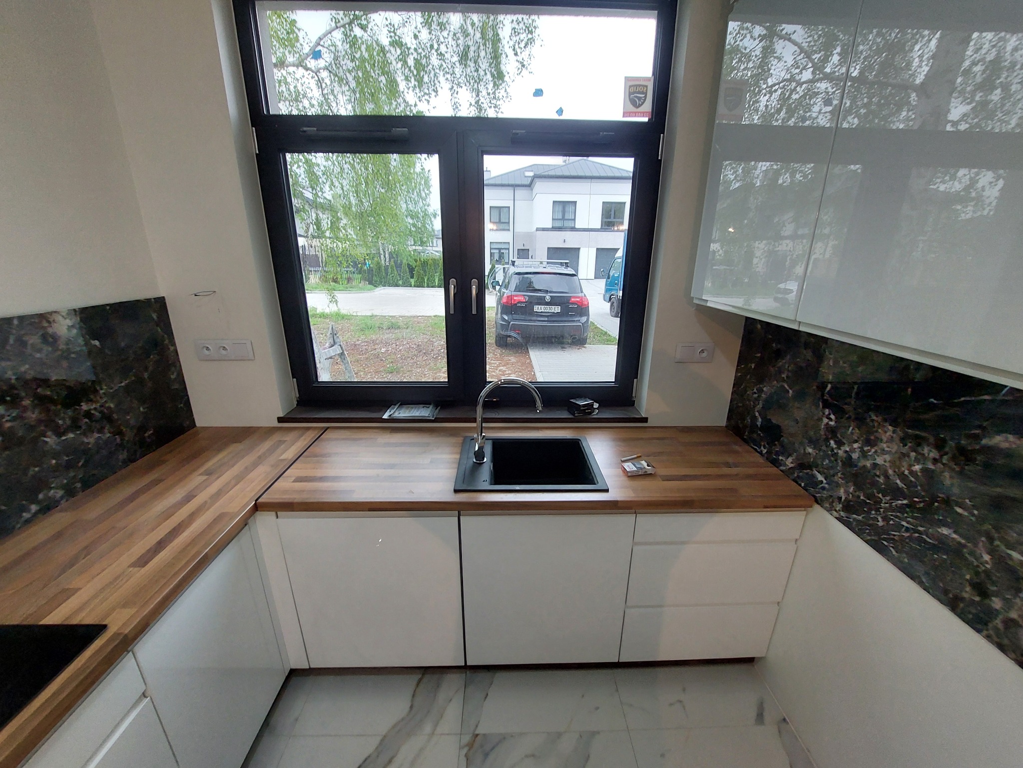 Nowoczesna kuchnia z białymi szafkami, drewnianym blatem i czarnym marmurem na ścianie, widok na podjazd z samochodem przez okno nad zlewem.