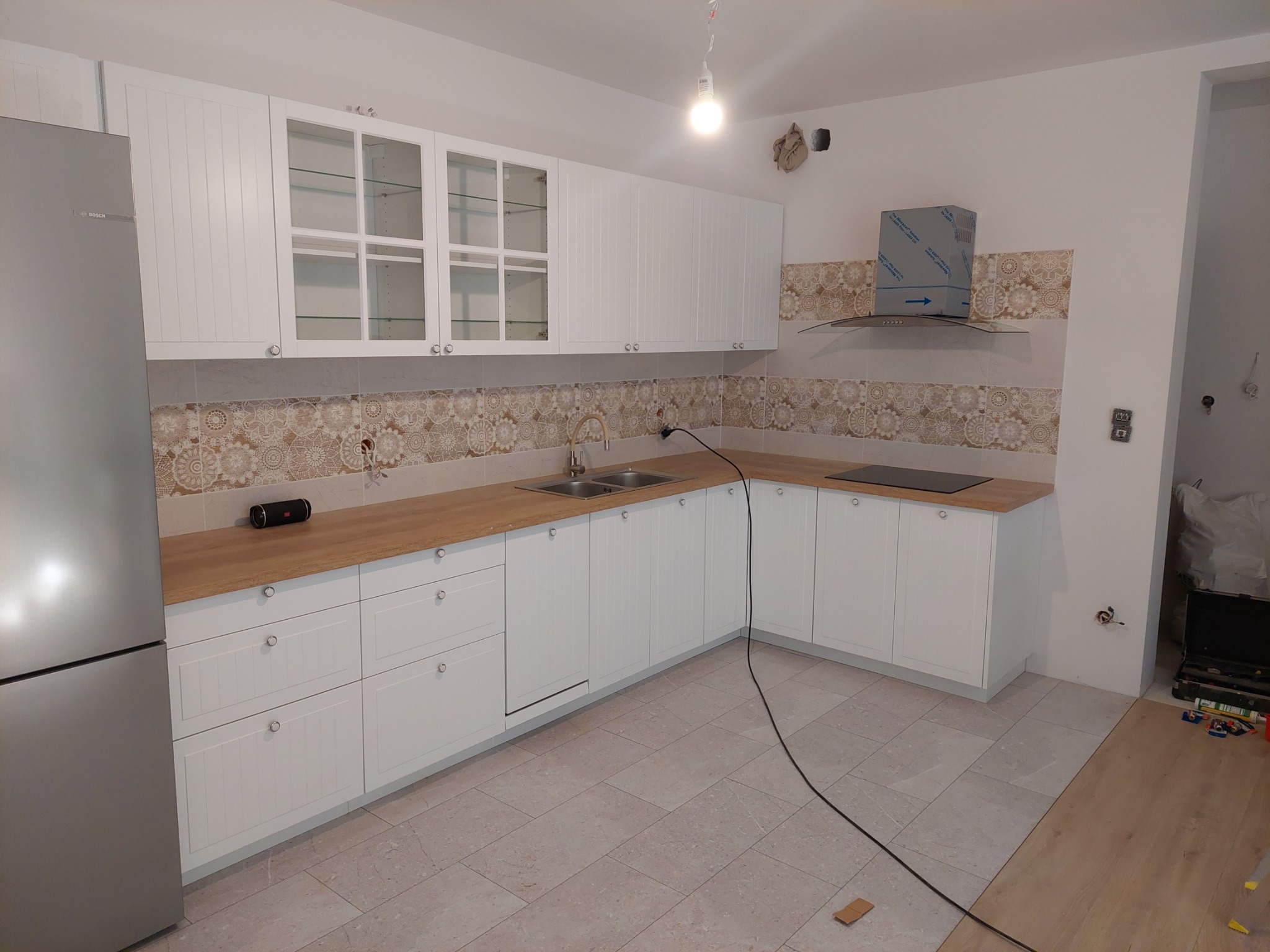 Biała zabudowa kuchenna z drewnianym blatem i szklanymi witrynami, widoczne elementy instalacji elektrycznej, płytki ceramiczne z wzorem i srebrna lodówka.