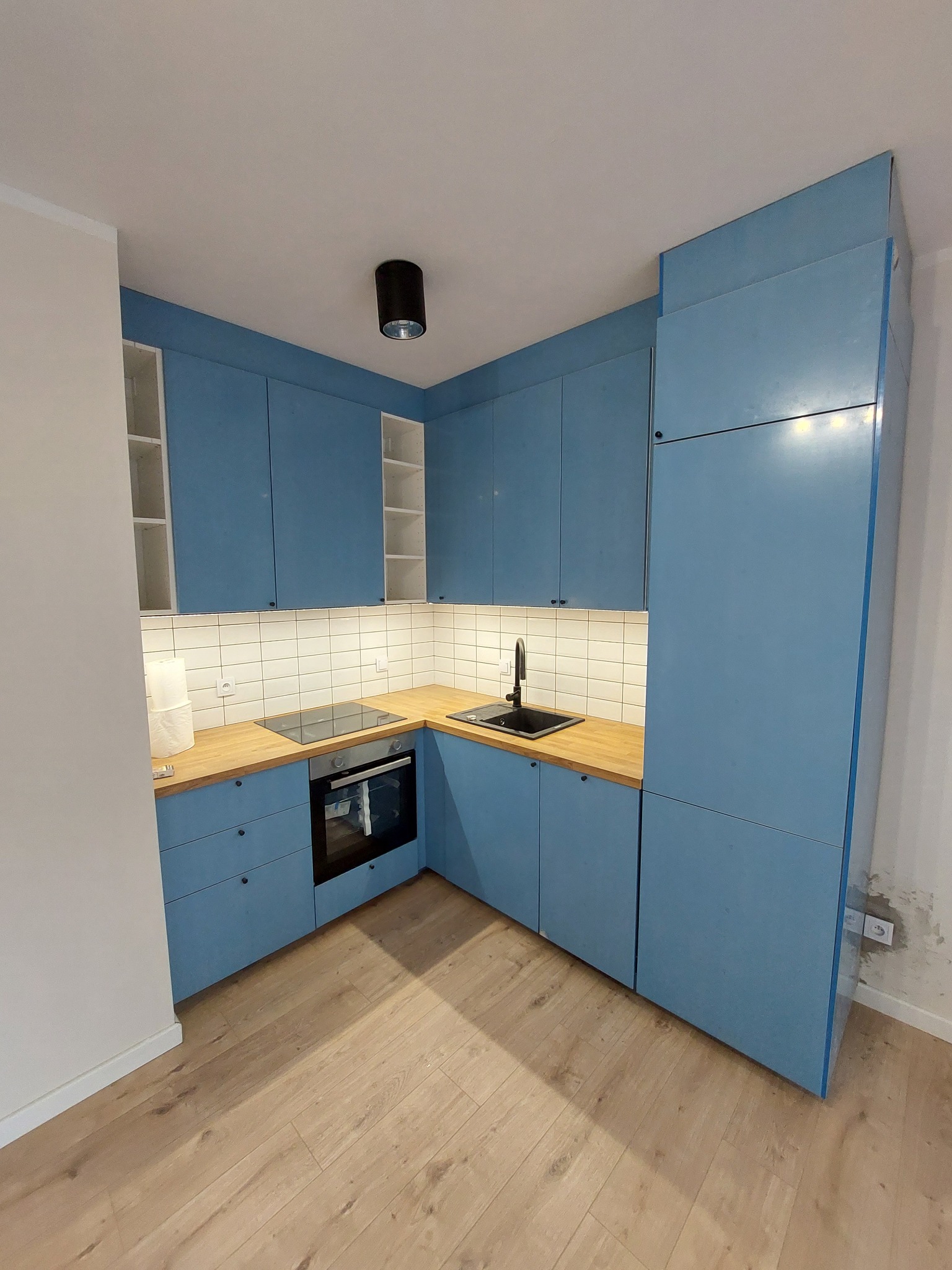 Niebieska, narożna kuchnia z drewnianym blatem, białymi płytkami i czarną armaturą, oświetlona punktowo, z widoczną płytą indukcyjną i piekarnikiem.