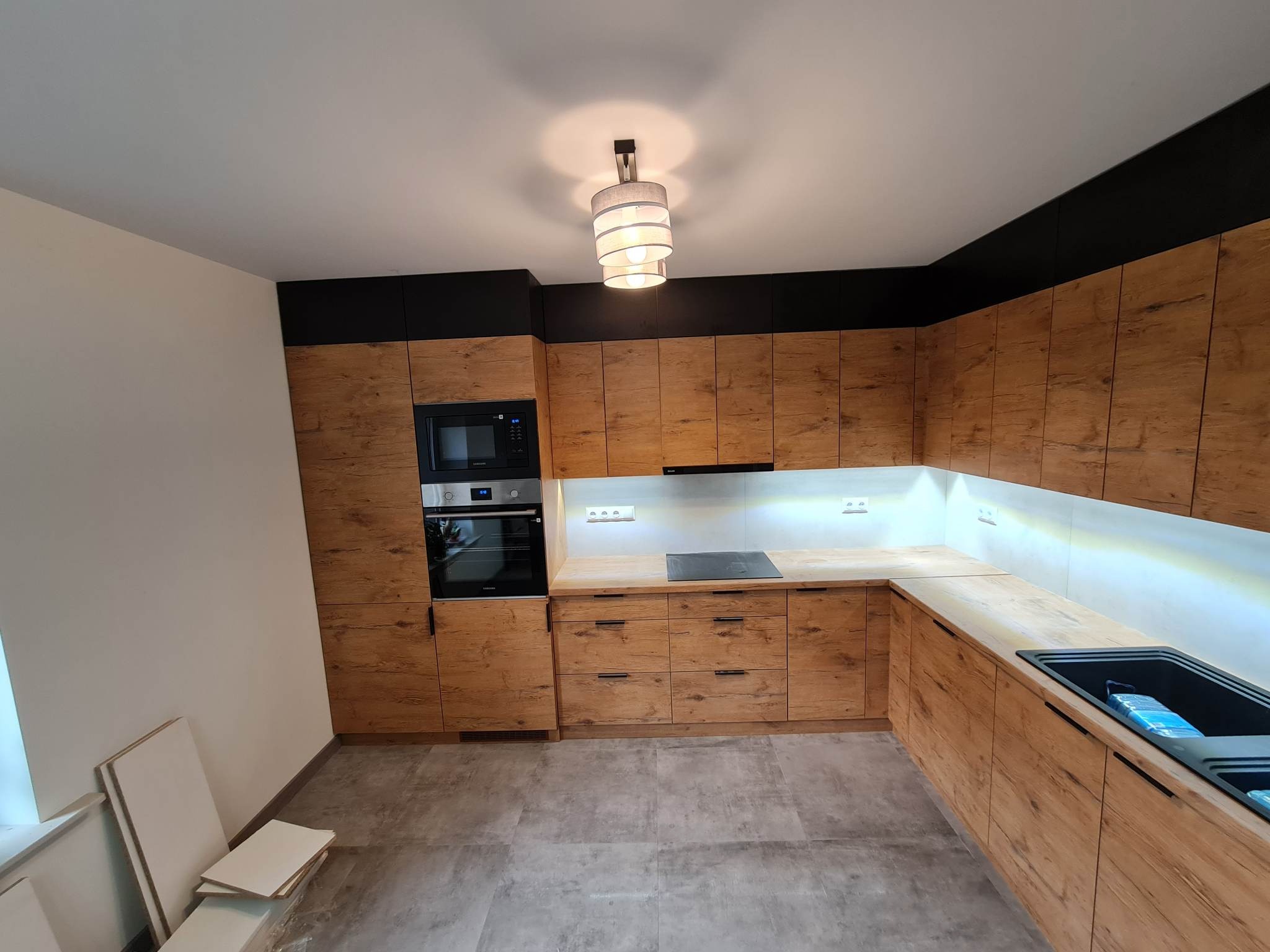 Nowoczesna kuchnia na wymiar z frontami w odcieniach drewna, czarne elementy dekoracyjne nad górnymi szafkami, oświetlenie LED pod szafkami wiszącymi i lampa sufitowa z abażurem.