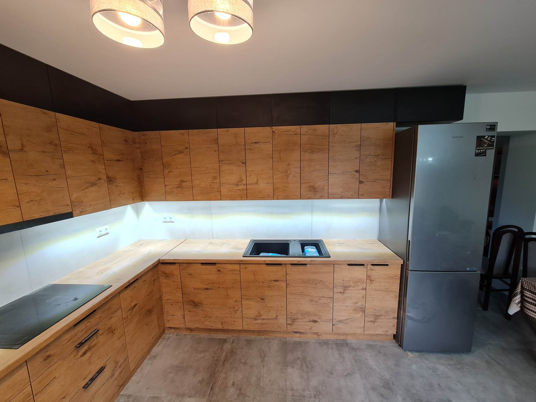 Nowoczesna kuchnia z drewnianymi szafkami o wyraźnej strukturze drewna, czarnymi górnymi szafkami, podświetleniem LED pod szafkami wiszącymi i szarą lodówką Samsung po prawej stronie.