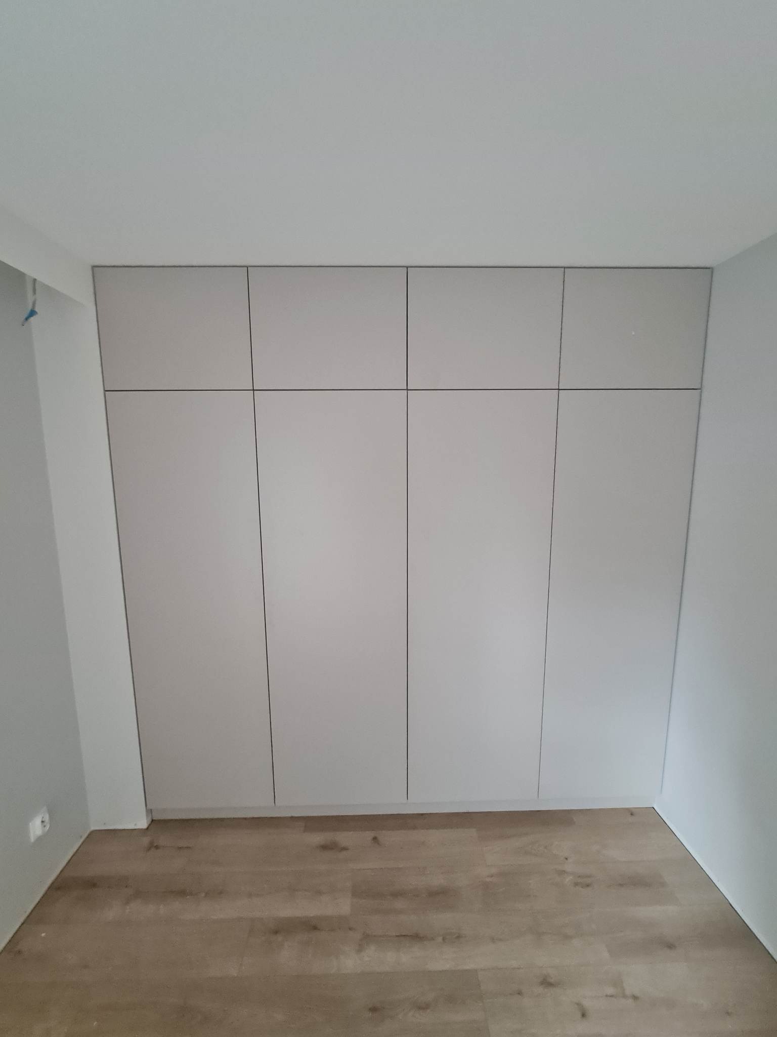 Jasnoszara szafa wnękowa z czterema drzwiami, dopasowana do sufitu, z widoczną podłogą z paneli w odcieniach brązu i szarości.