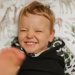 DK między kadrami - Uśmiechnięty chłopiec z blond włosami, leżący na wzorzystej pościeli z motywem zwierząt, z zaciśniętymi oczami i widocznymi zębami, ubrany w czarną bluzę z kapturem w biało-zielone wzory. W kadrze...