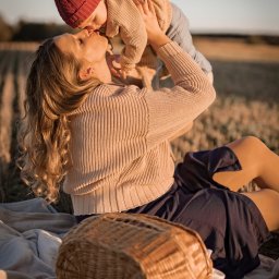 DK między kadrami - Matka w beżowym swetrze siedzi na kocu w polu, podnosząc i całując małe dziecko w czerwonej czapce z pomponem, obok koszyk piknikowy.