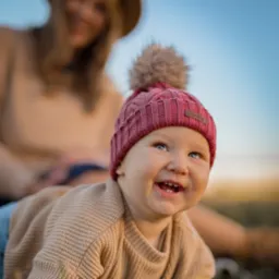 Uśmiechnięte niemowlę w bordowej czapce z pomponem, leżące na brzuszku, z zarysowaną sylwetką matki w tle.