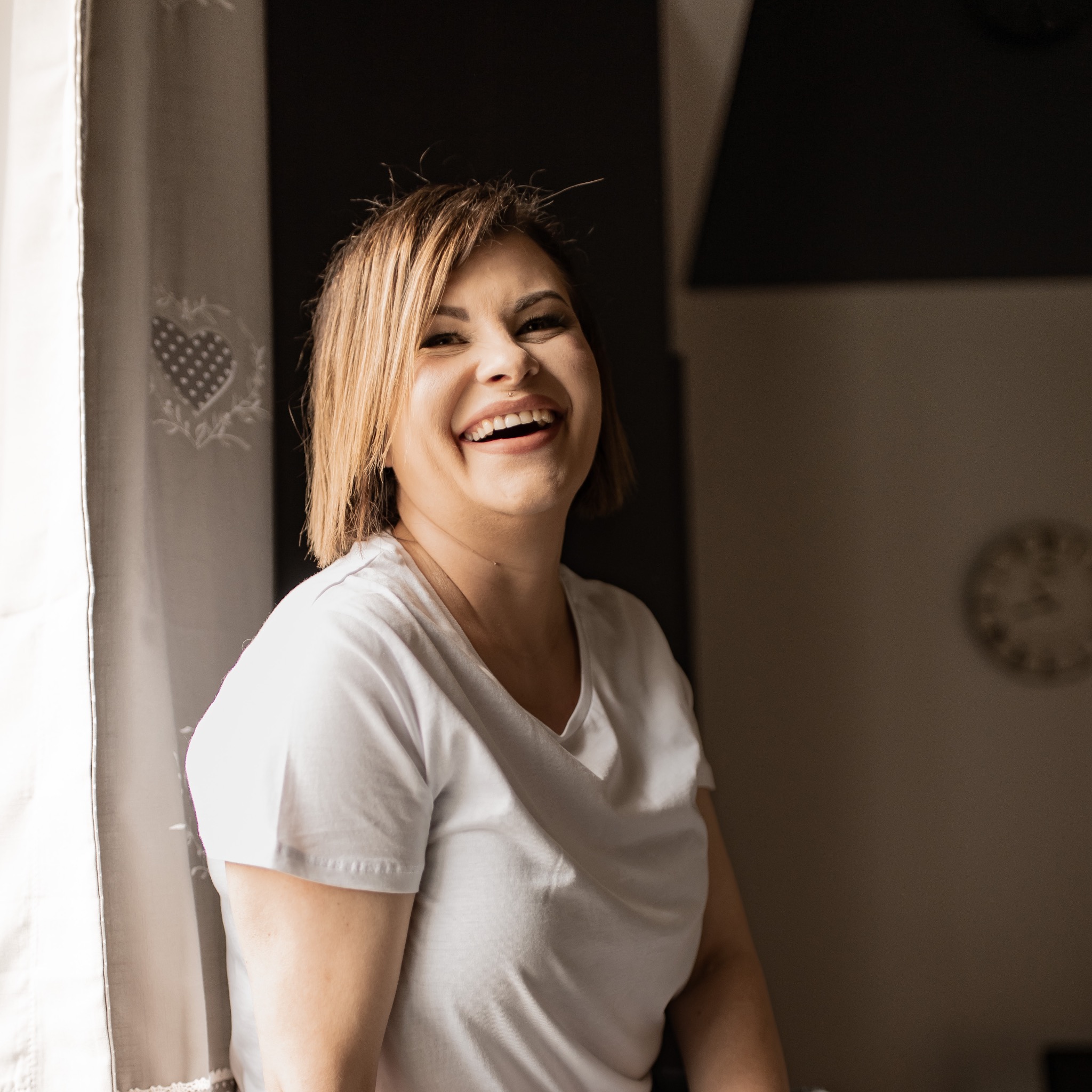 Portret roześmianej kobiety w białej koszulce, stojącej przy oknie z zasłoną w serduszka, z widocznym zegarem na ścianie w tle.