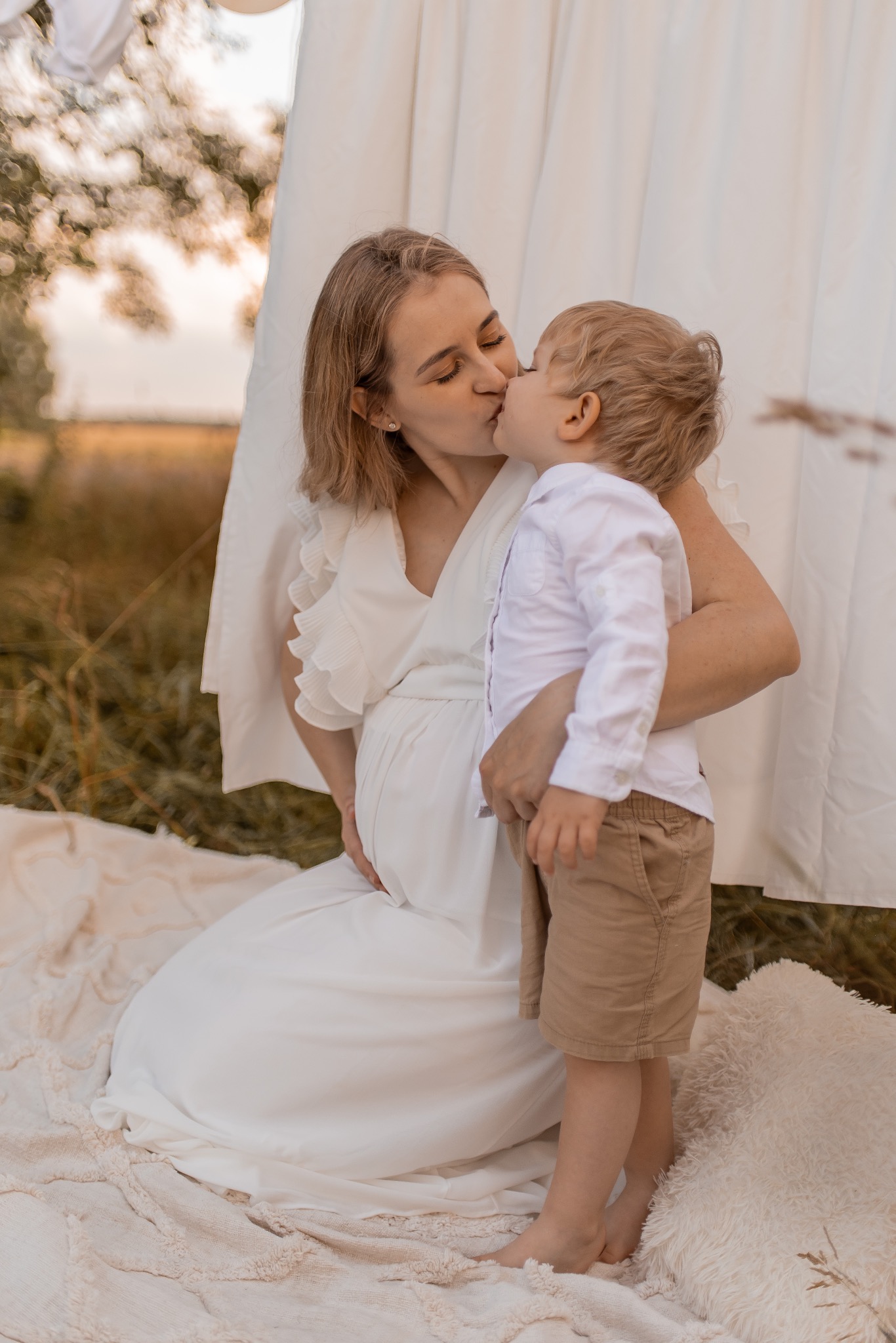 Ciężarna kobieta w białej sukience klęczy na kocu, tuląc i całując małego chłopca w beżowych szortach na tle białej tkaniny i polnej roślinności.