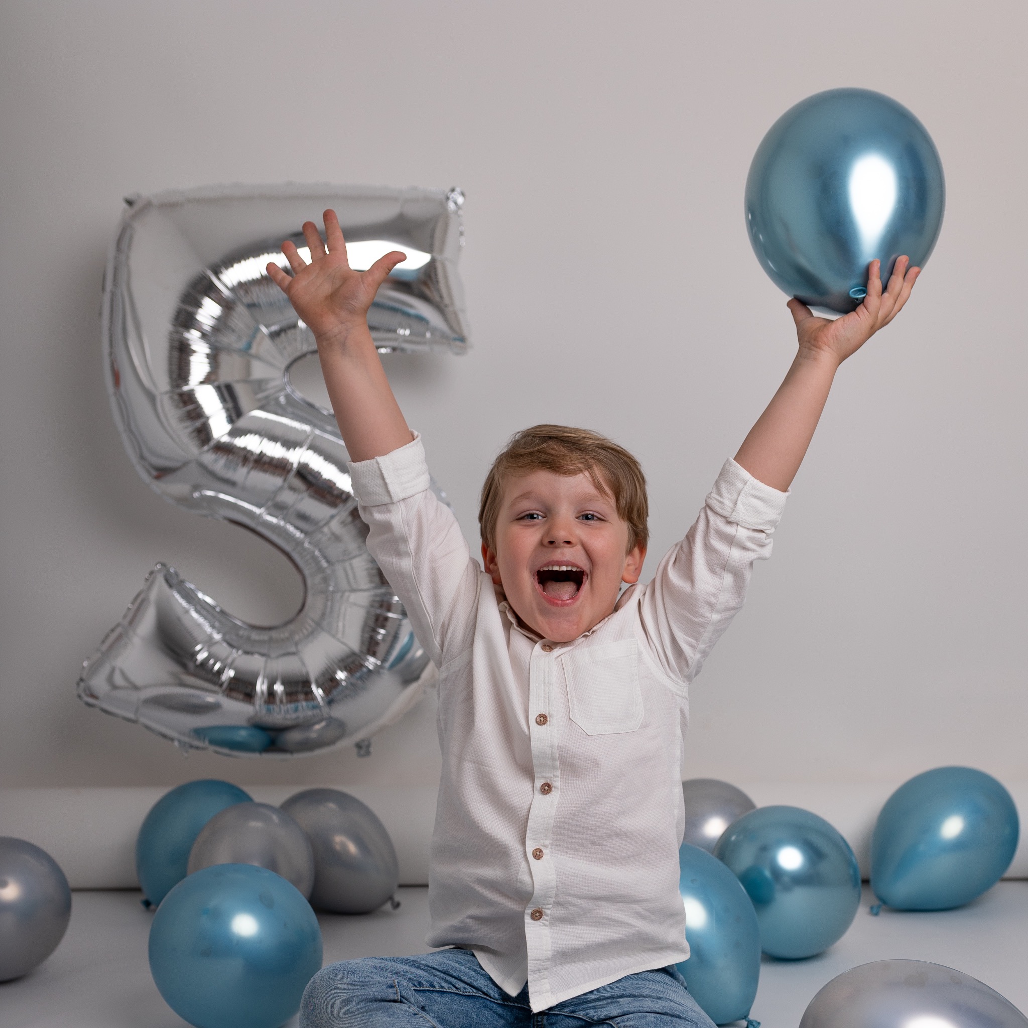 Rozradowany chłopiec z blond włosami siedzi na podłodze z balonami w odcieniach srebra i błękitu, trzymając jeden z nich w górze, obok niego duży balon foliowy w kształcie cyfry 5.