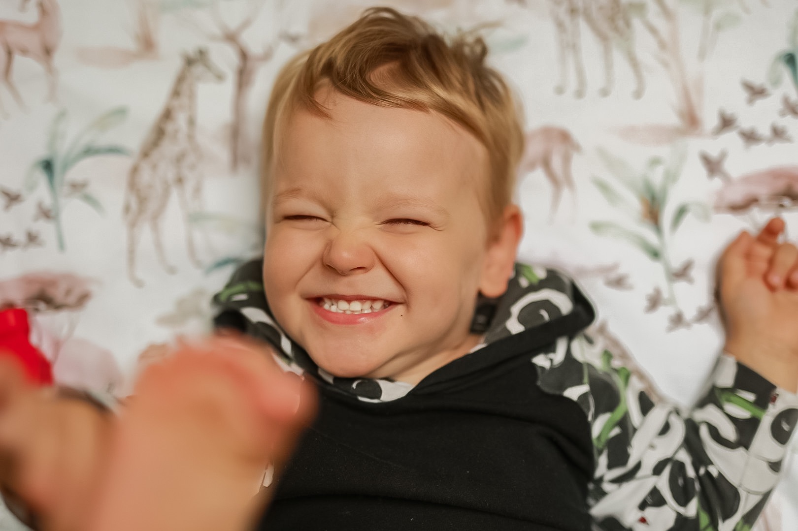 Uśmiechnięty chłopiec z blond włosami, leżący na wzorzystej pościeli z motywem zwierząt, z zaciśniętymi oczami i widocznymi zębami, ubrany w czarną bluzę z kapturem w biało-zielone wzory. W kadrze...