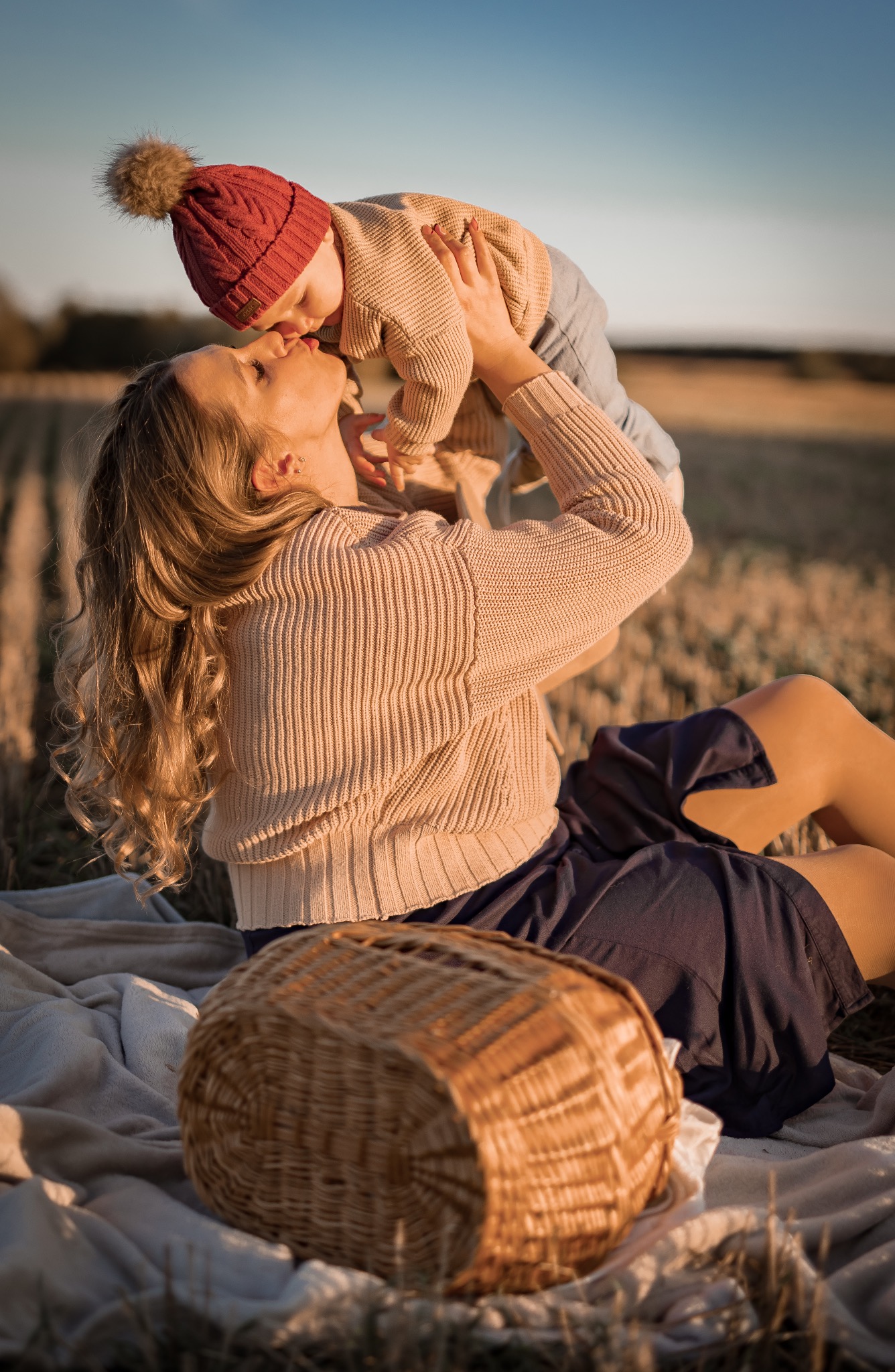 Matka w beżowym swetrze siedzi na kocu w polu, podnosząc i całując małe dziecko w czerwonej czapce z pomponem, obok koszyk piknikowy.