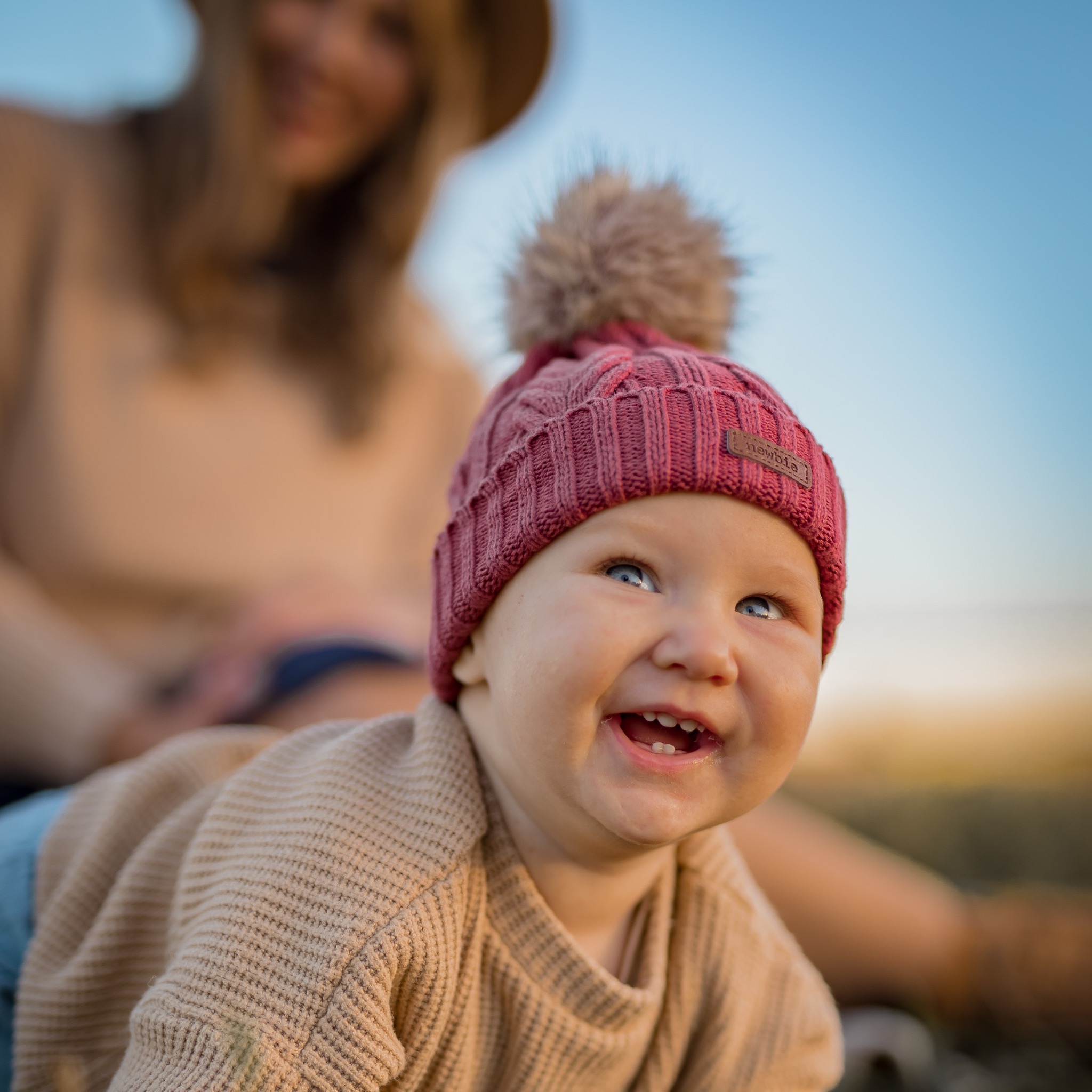 Uśmiechnięte niemowlę w bordowej czapce z pomponem, leżące na brzuszku, z zarysowaną sylwetką matki w tle.