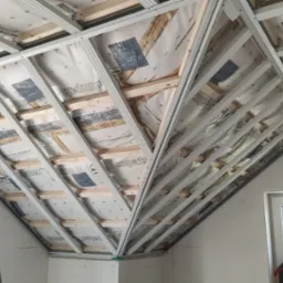 Skośny sufit poddasza w trakcie montażu: widoczna konstrukcja z profili, izolacja z folii aluminiowej. Okno z prawej strony, drabina w lewym dolnym rogu.