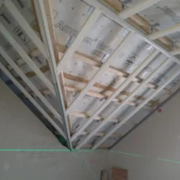 Szkielet drewniany poddasza z izolacją, widoczne elementy konstrukcyjne i laserowy wskaźnik poziomu. Prace przygotowawcze przed montażem płyt.