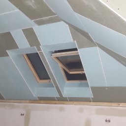 Robert Grzywinski - Skomplikowana konstrukcja sufitu poddasza z oknami dachowymi, częściowo pokryta płytami gipsowo-kartonowymi, widoczne łączenia i szpachlowanie.