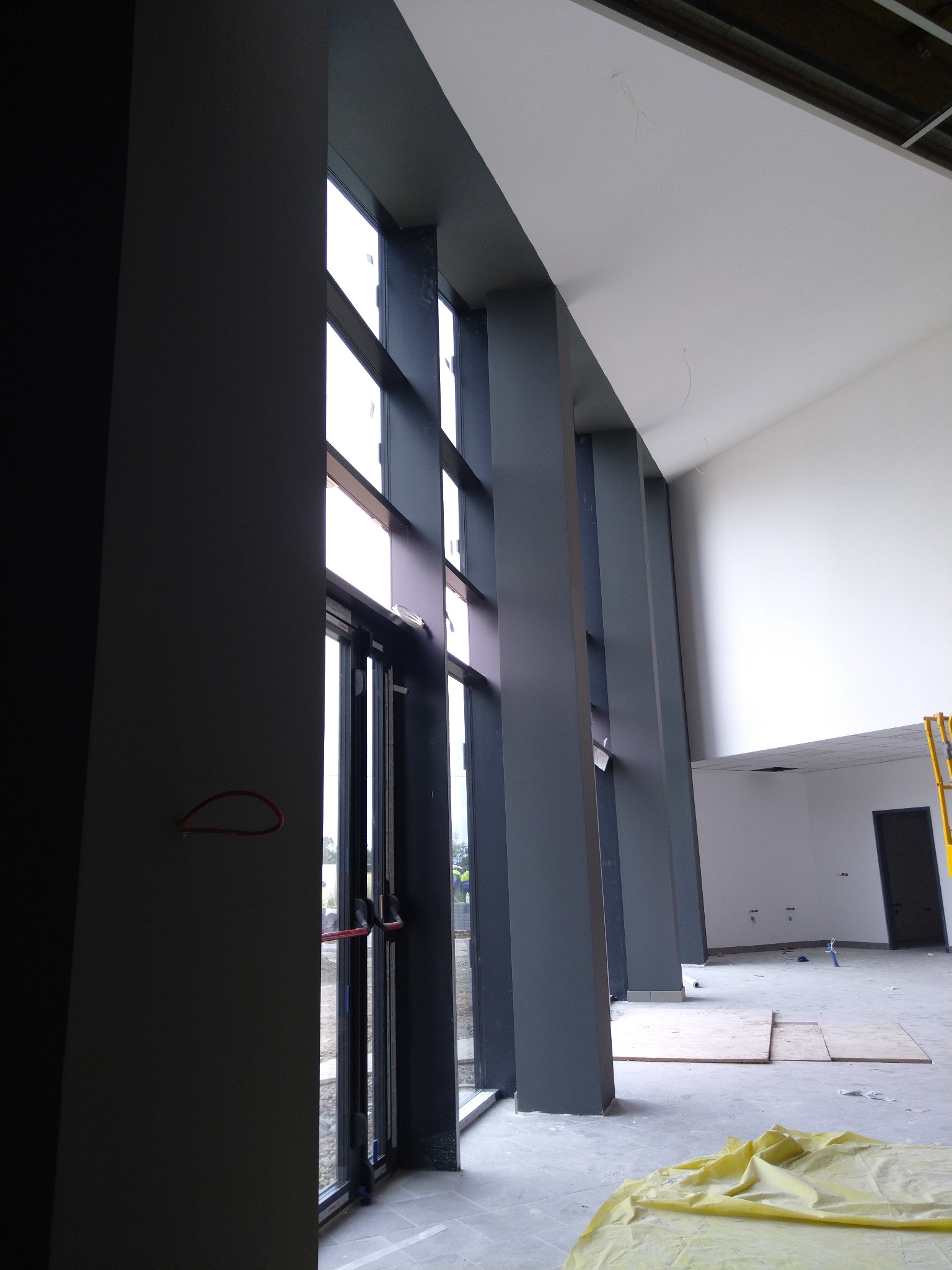 Wnętrze w trakcie remontu z wysokimi oknami i kolumnami, widoczna surowa ściana i zabezpieczona podłoga.